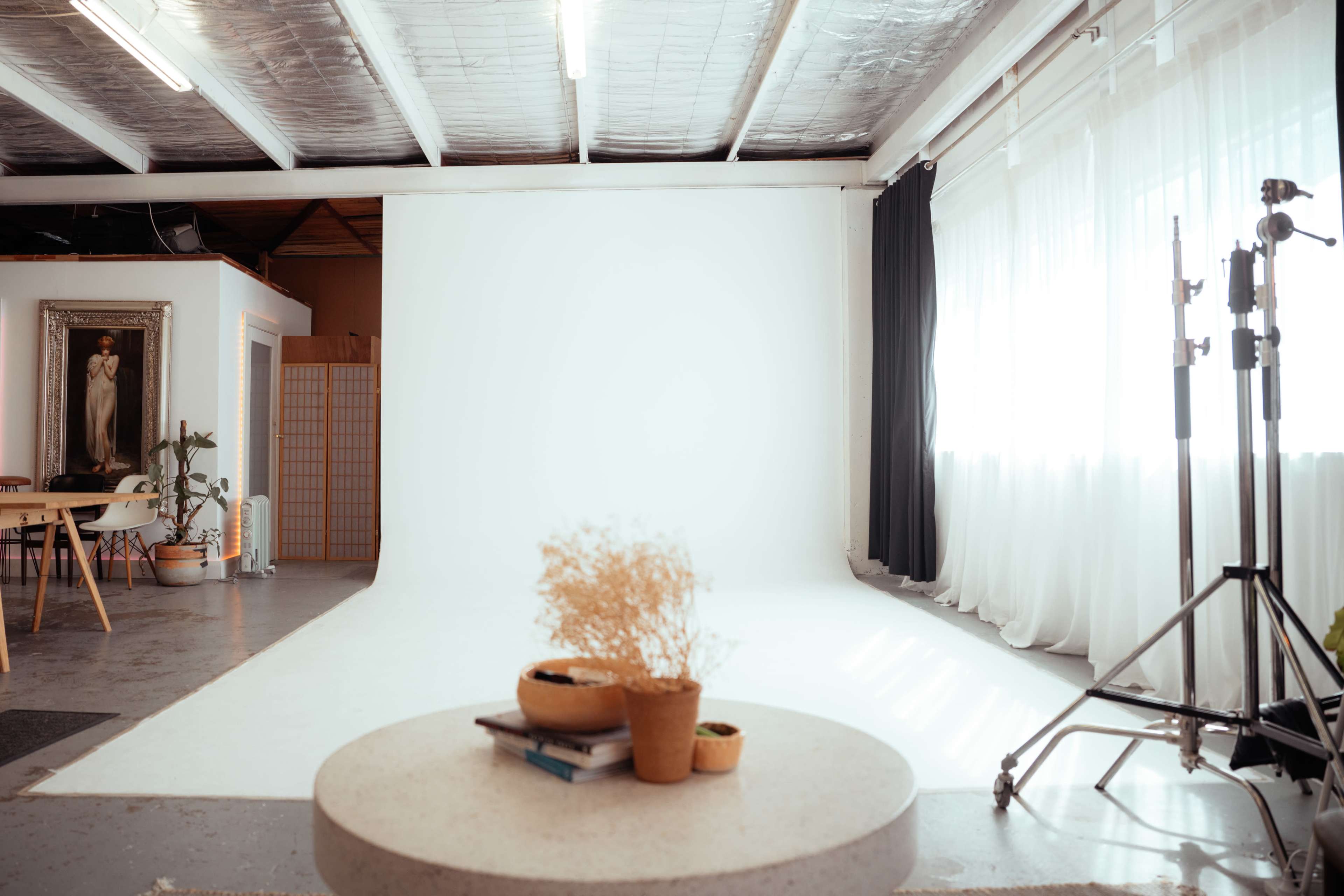 The image shows a photography studio with a large white backdrop, a round coffee table with decorations in the center, and natural light coming through large windows.