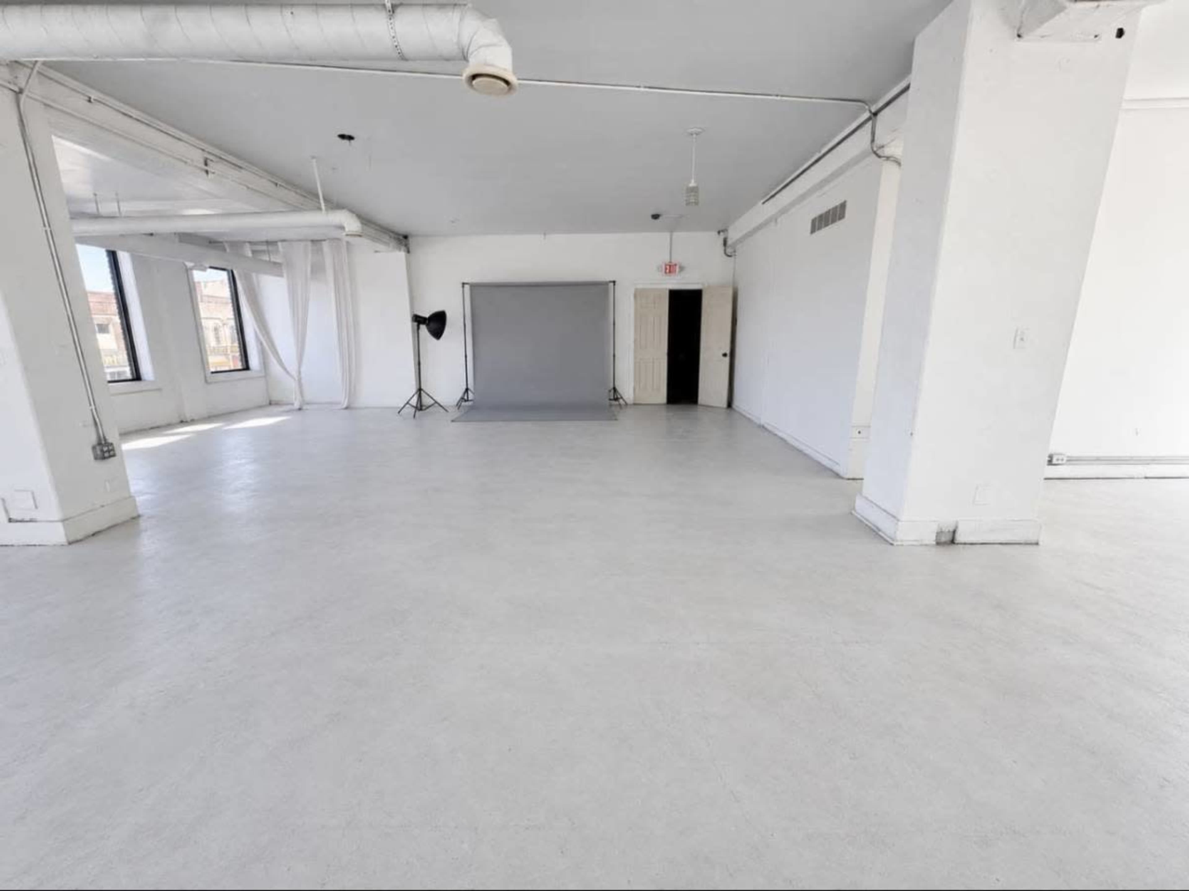 The image shows a spacious, empty room with white walls, a grey backdrop on a stand, and large windows allowing natural light.