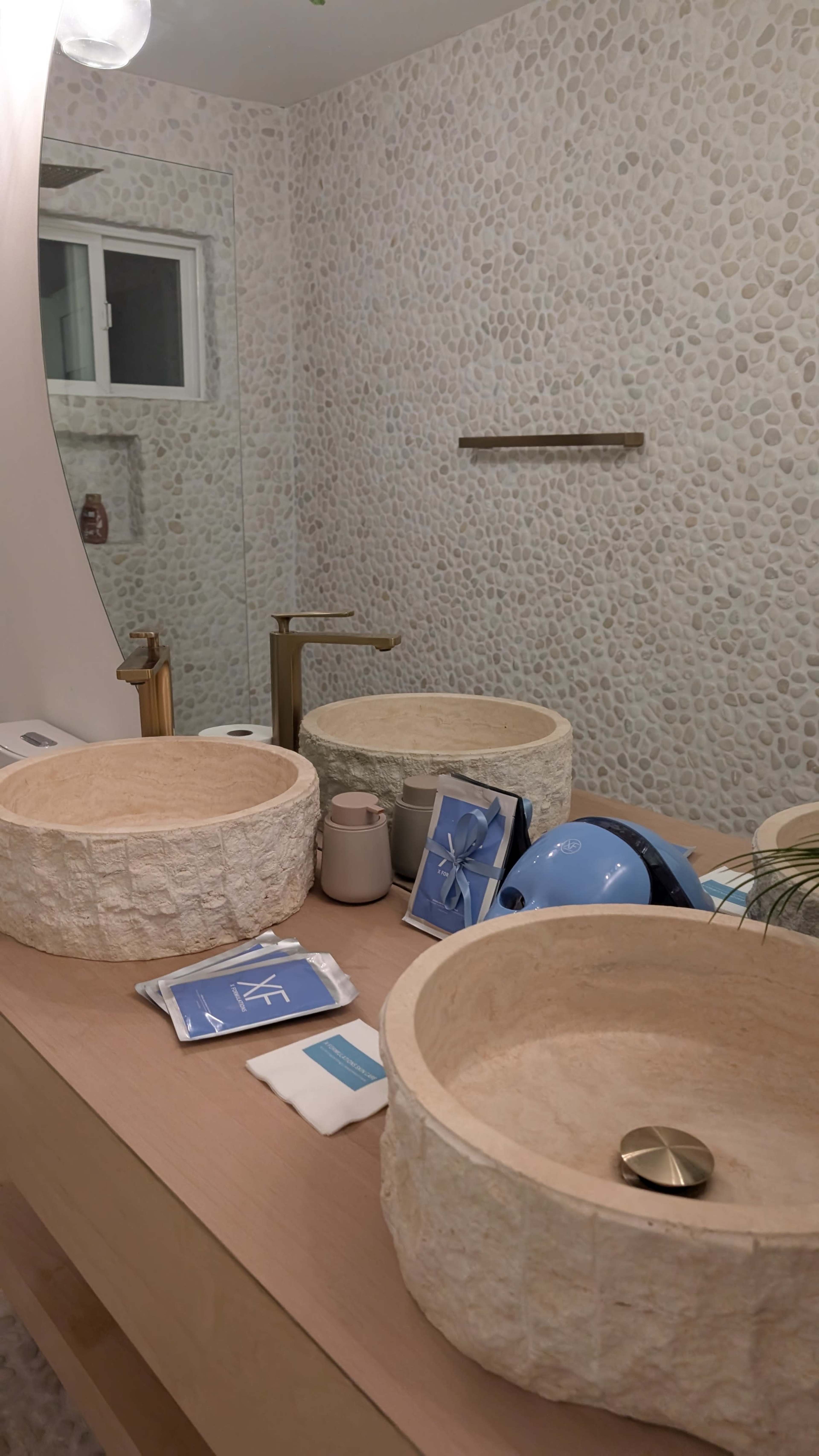 The image depicts a modern bathroom featuring two circular stone sinks on a wooden vanity, with various toiletries and decorative items arranged nearby.