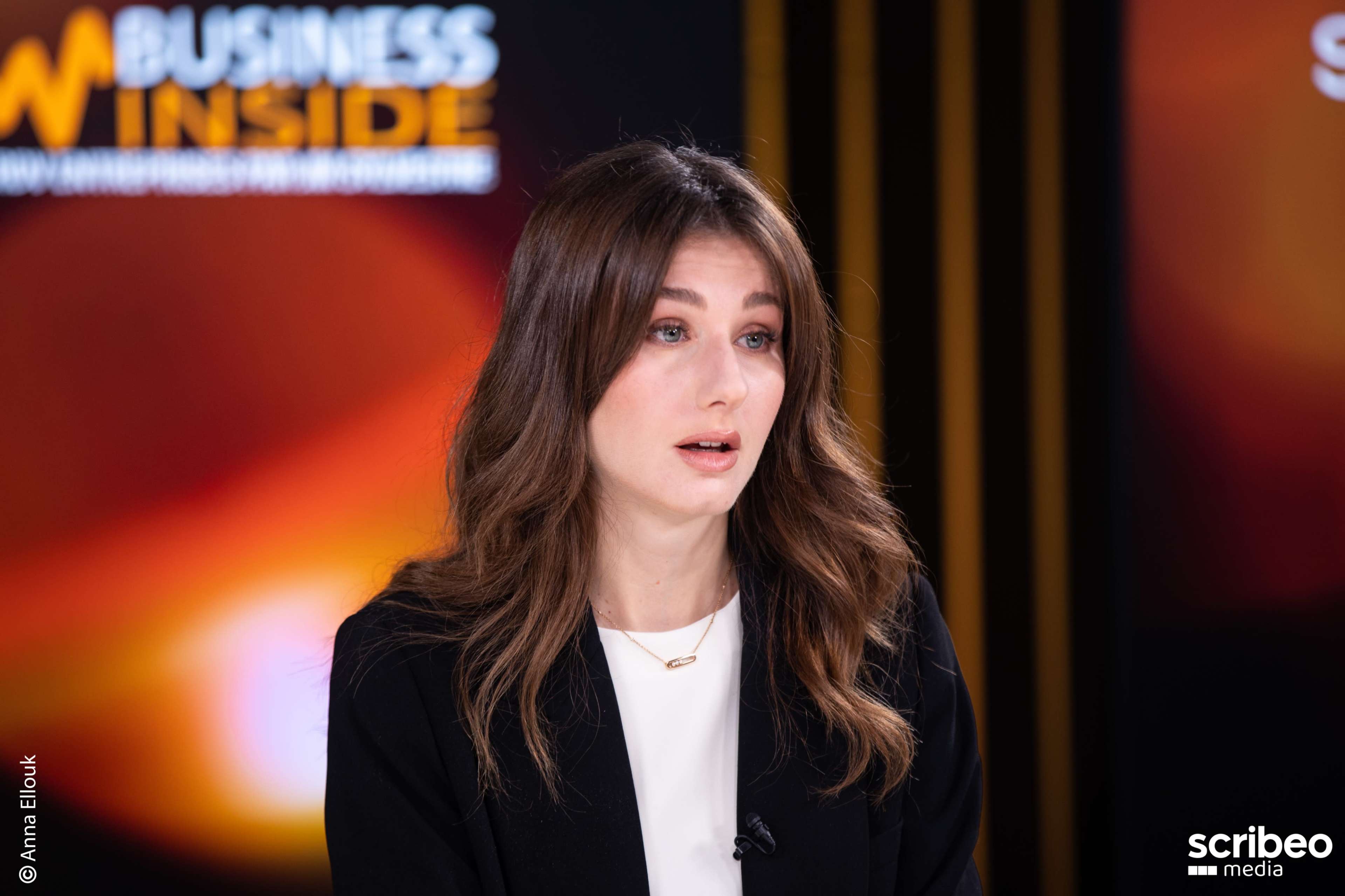 A woman with long, brown hair is seated in front of a brightly lit backdrop, speaking into a microphone during an interview.