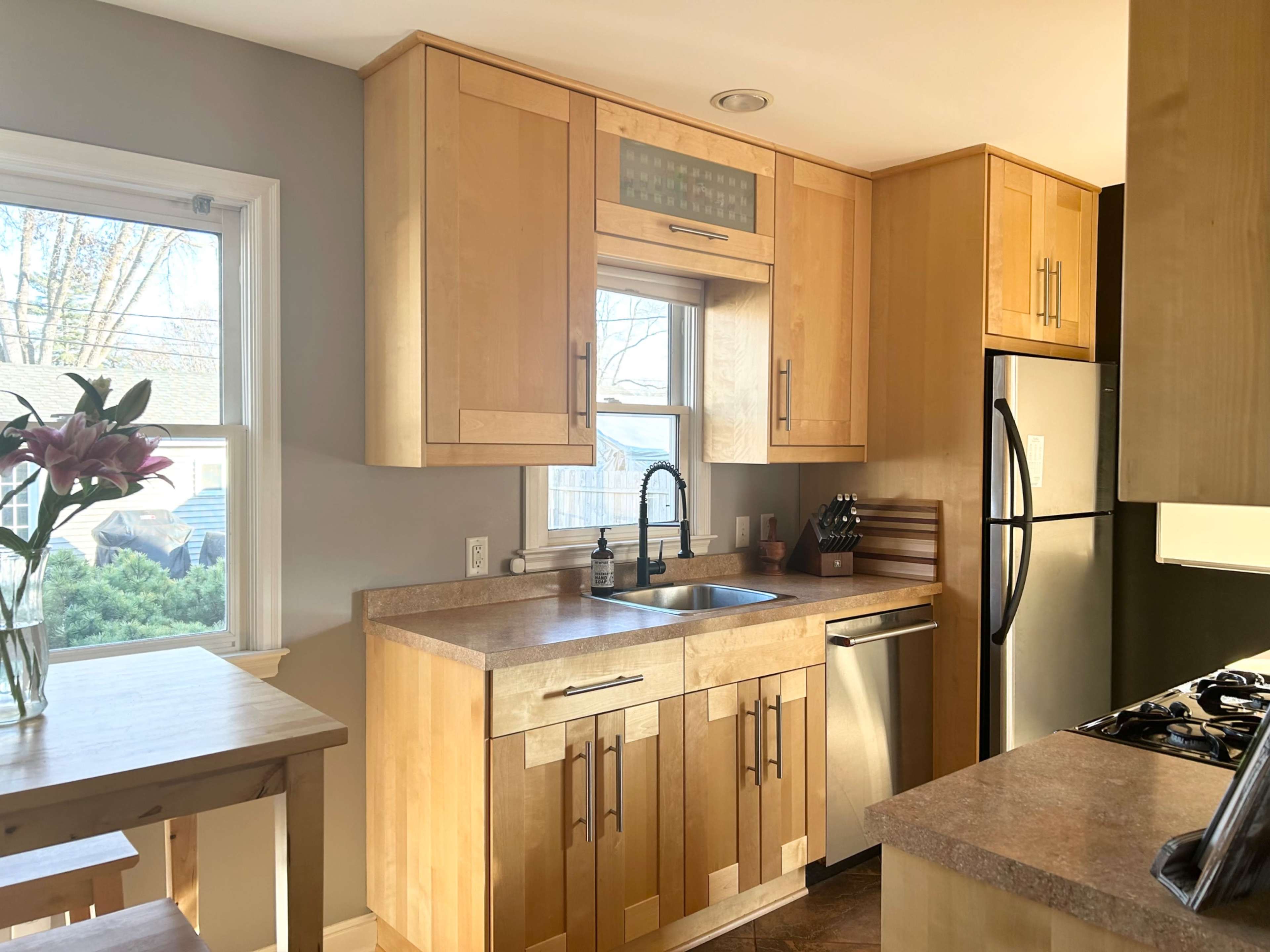 The image shows a modern kitchen with wooden cabinetry, a stainless steel refrigerator, and a window above the sink.