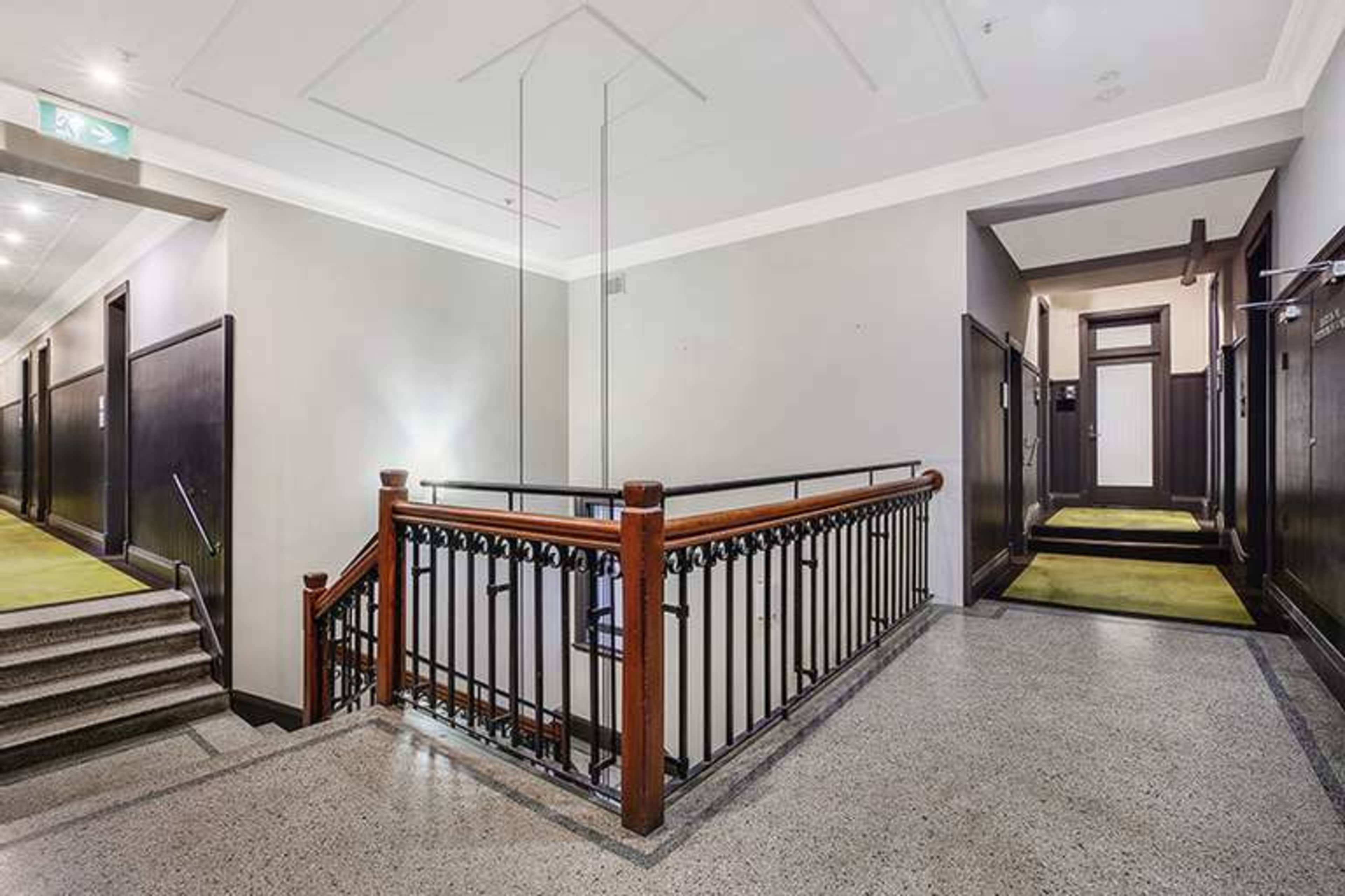 A well-lit staircase with a wooden railing, leading to a landing area that connects to several closed doors along the corridor.