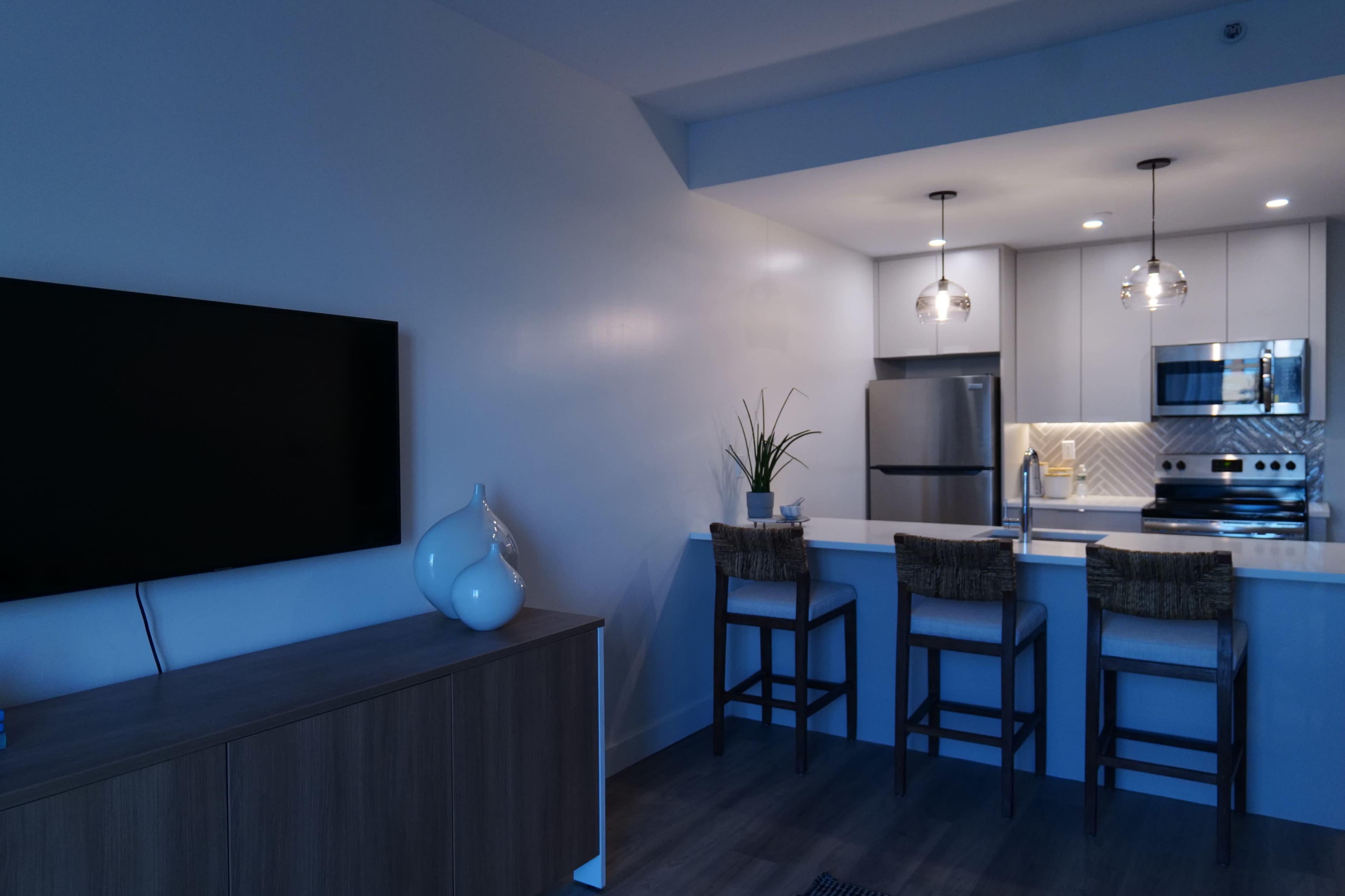 A modern kitchen and living area with a television on a cabinet, bar stools at a kitchen counter, and stainless steel appliances.