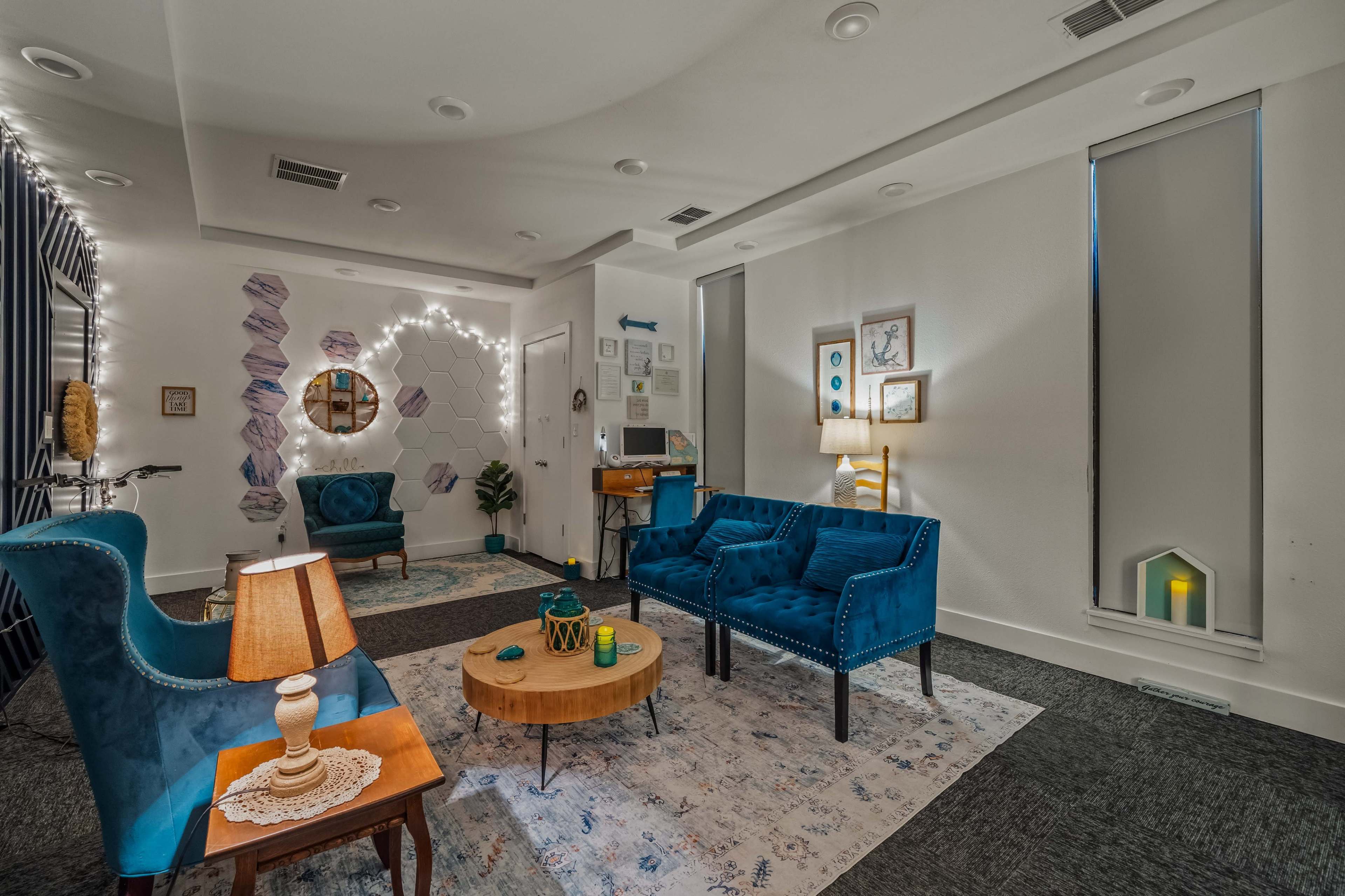 The scene features a cozy lounge area with blue upholstered furniture, a round wooden coffee table, decorative wall art, and soft lighting.