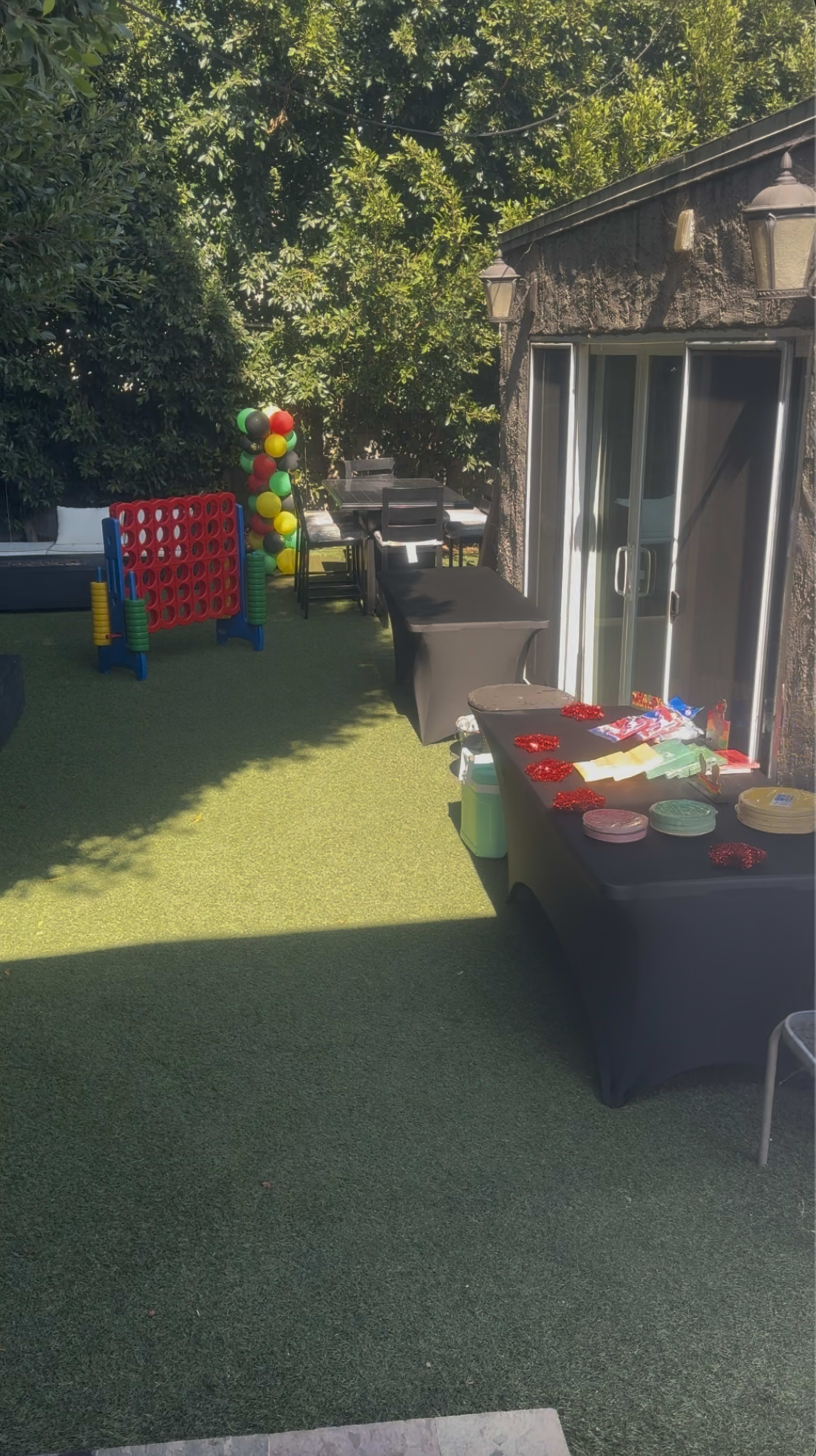 The image shows a backyard party setup with tables covered in black cloth, colorful decorations, and games, including a large Connect Four set.