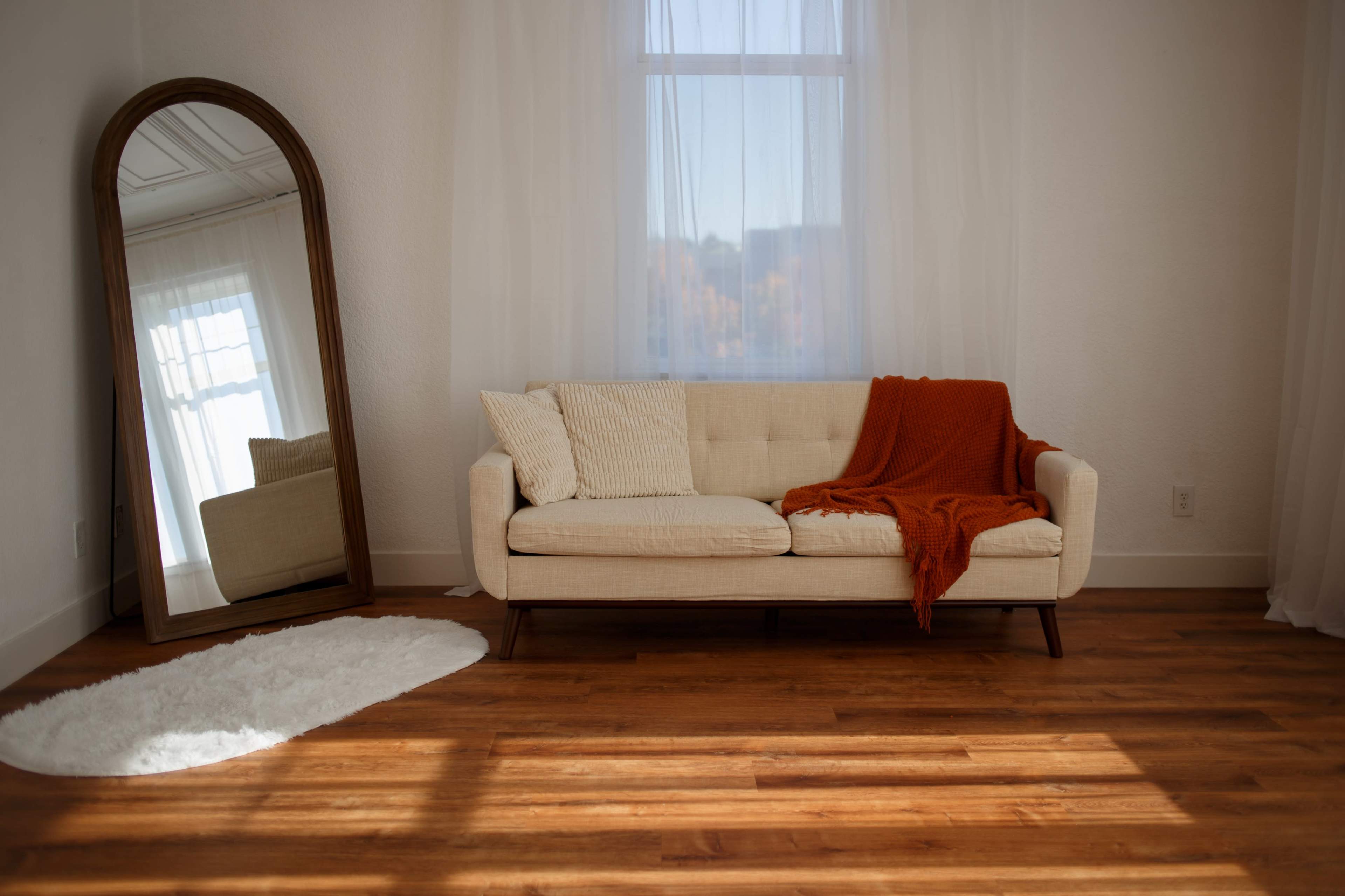 A light-colored sofa with decorative pillows sits in a bright room beside a large mirror and a round rug on a wooden floor.