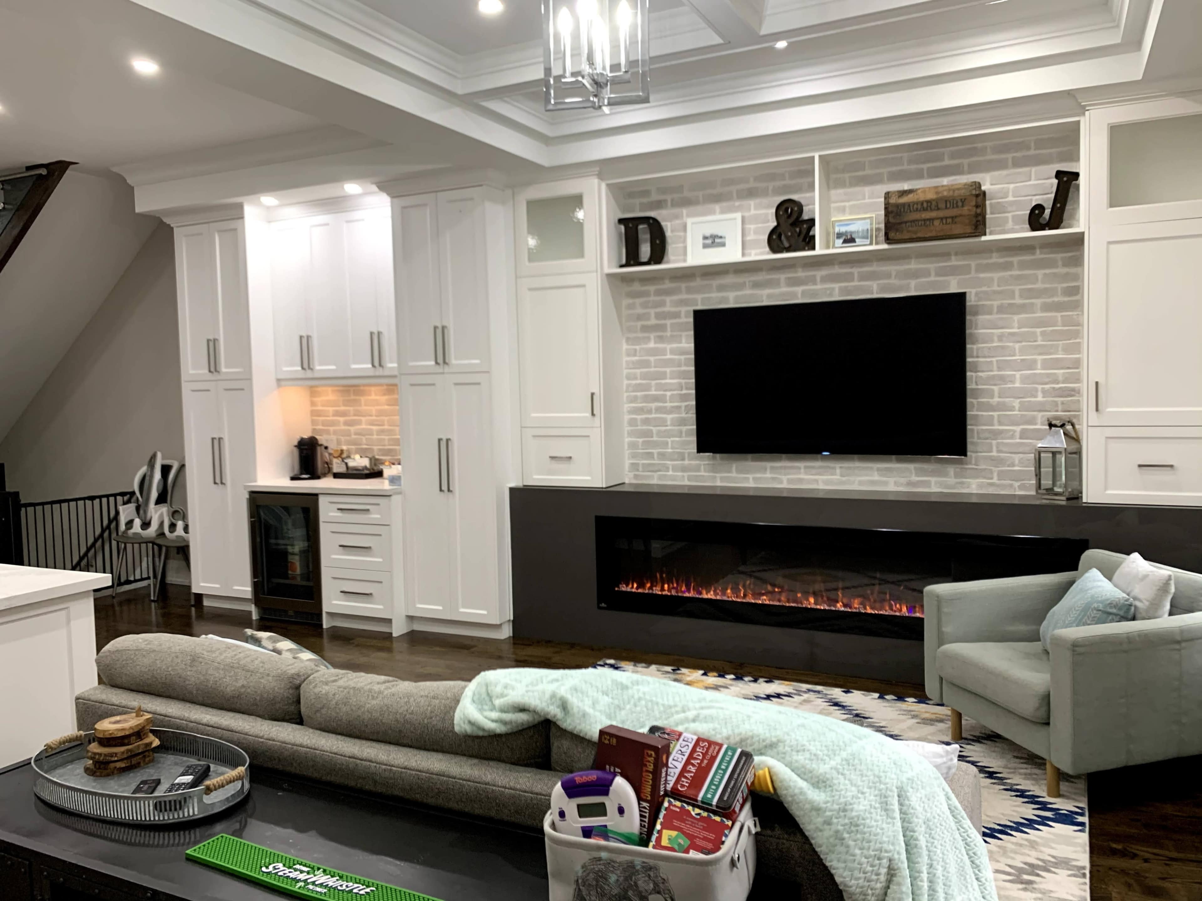 A modern living room with a gray sofa, a wall-mounted TV above a fireplace, and a white kitchen area in the background.