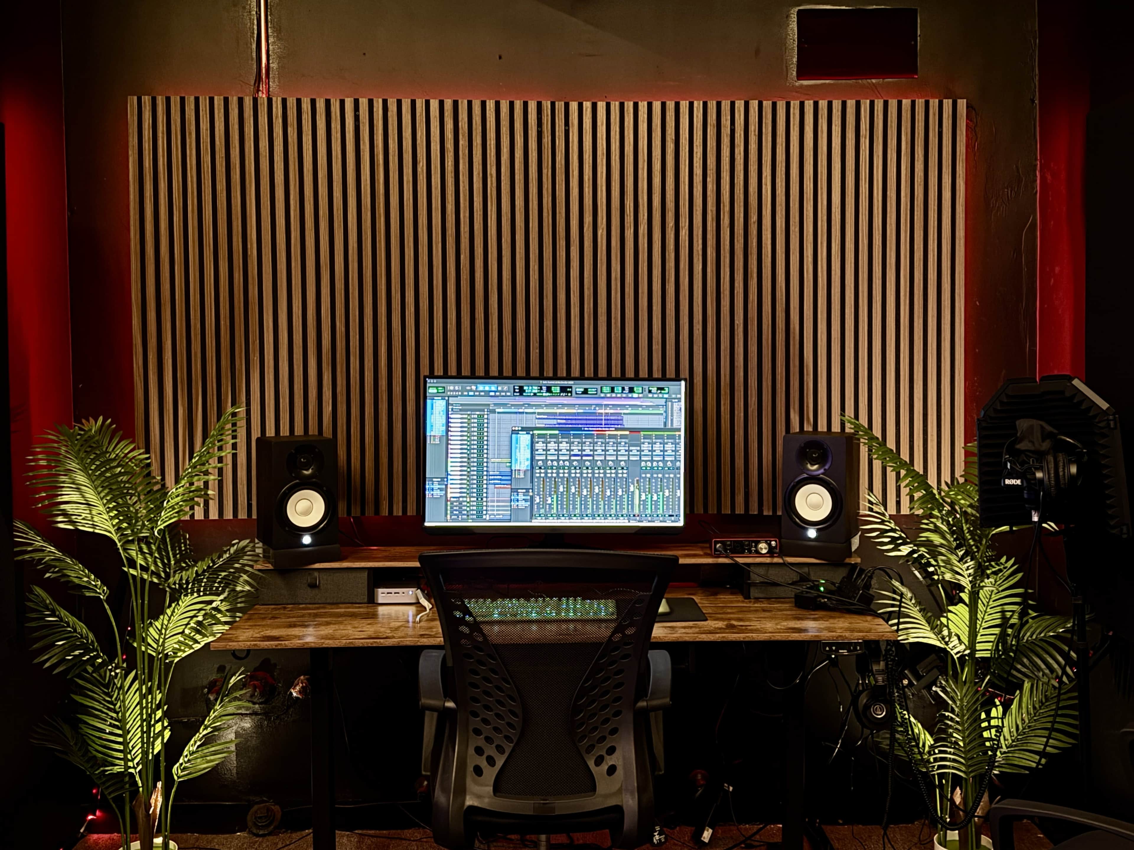 The image shows a music production studio with a computer monitor displaying audio software, two speakers on either side, and plants flanking the desk.
