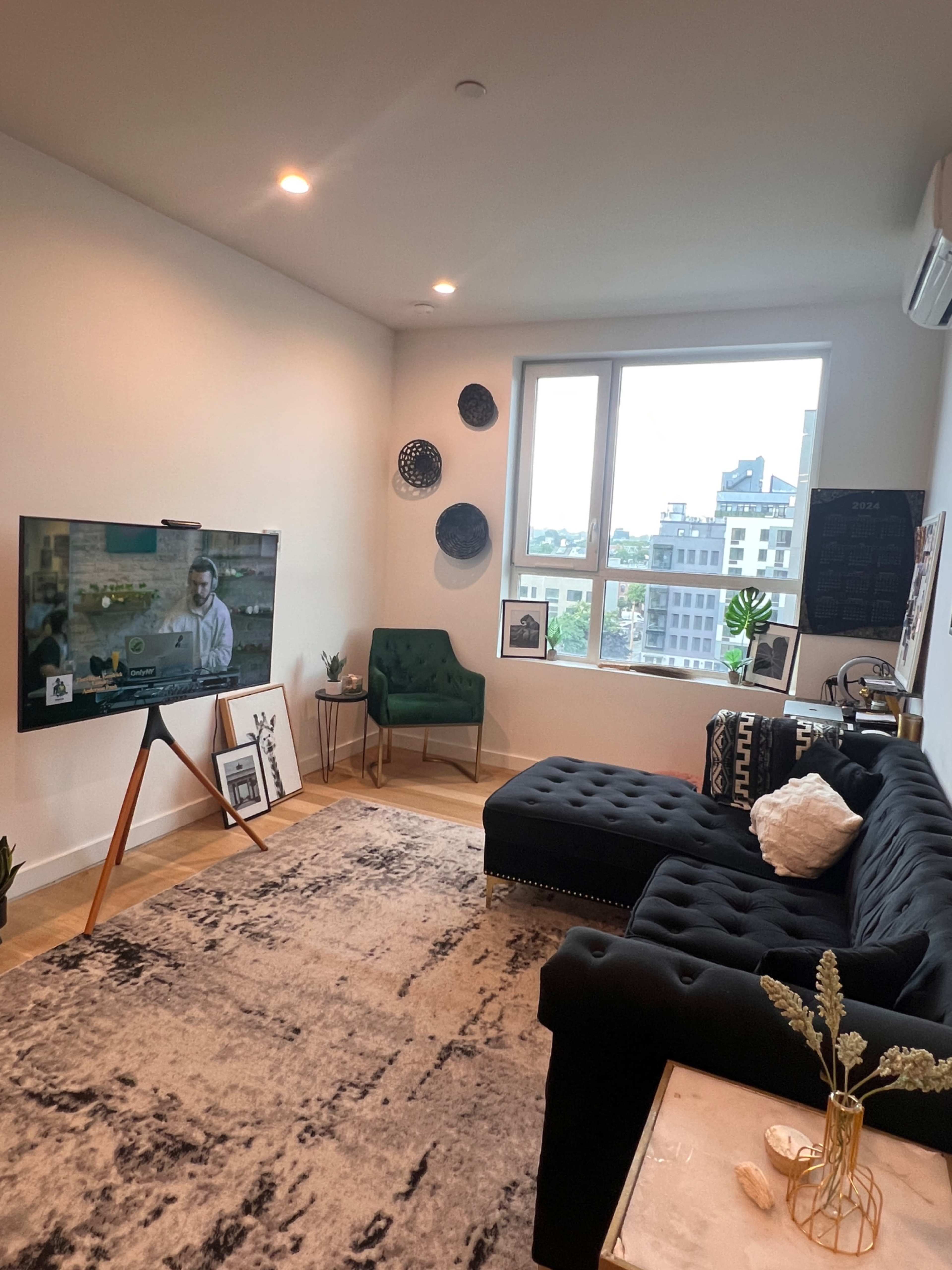 A modern living room with a black tufted sectional sofa, a TV on a wooden stand, and a large window overlooking an urban landscape.