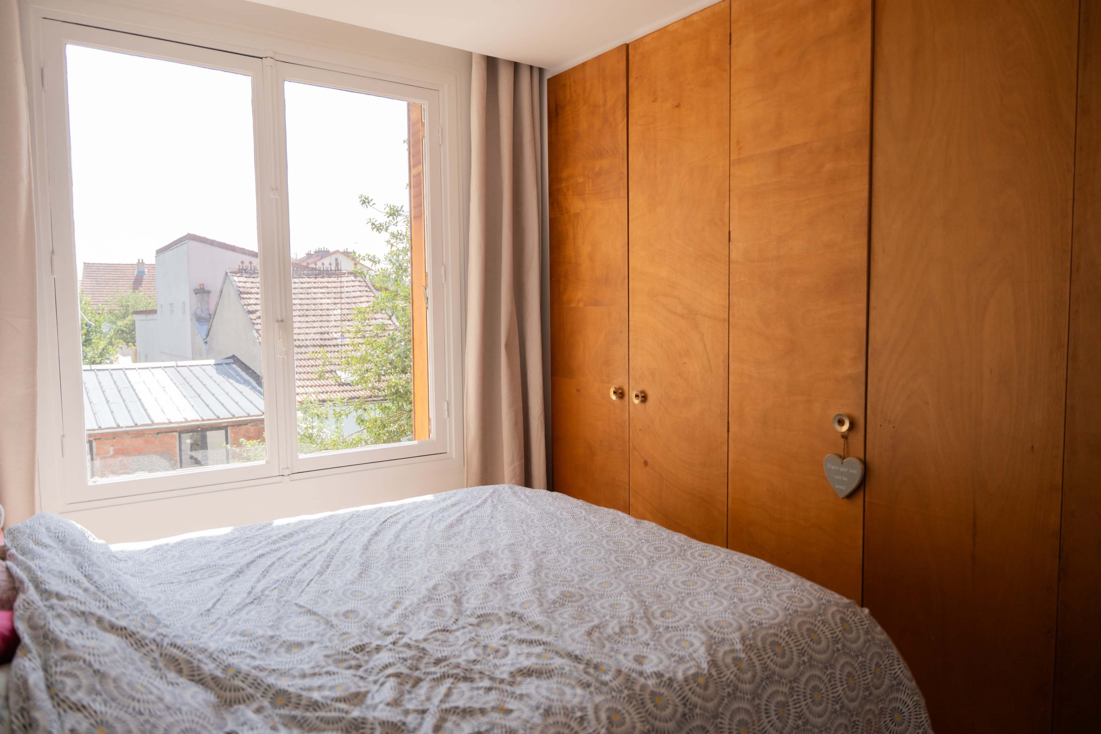 A bedroom features a light-colored bedspread in front of a window overlooking a residential area, with wooden cabinetry on one side.