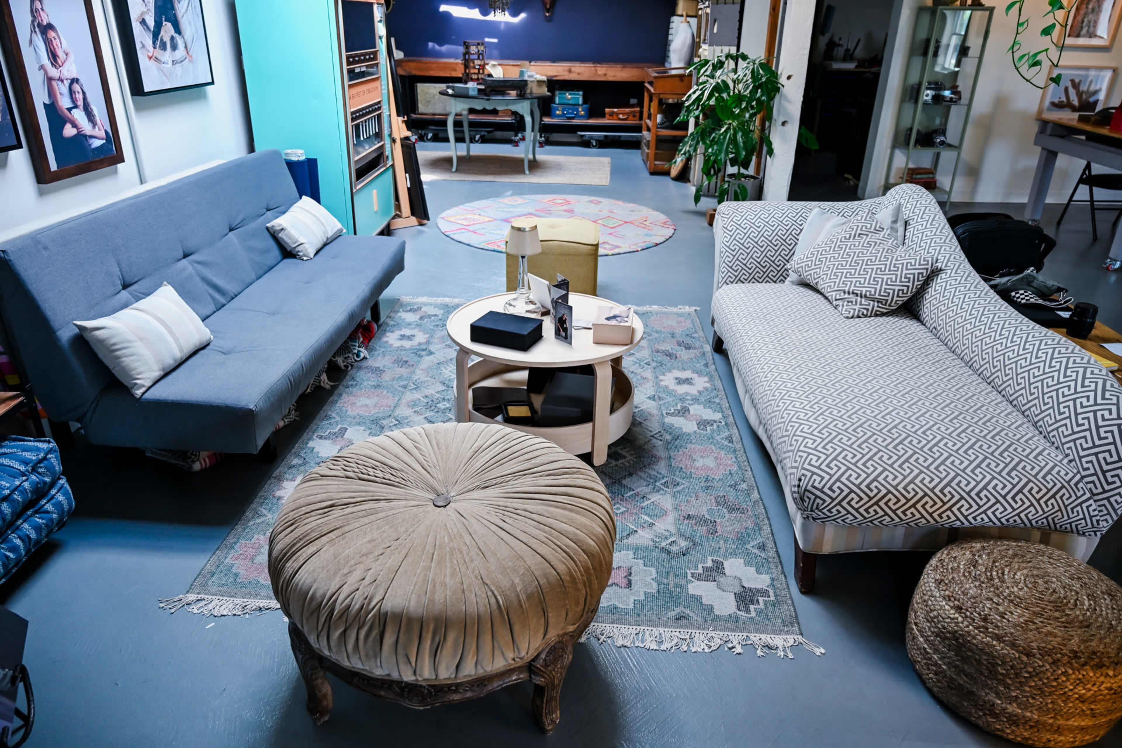 The image shows a cozy living room featuring a gray futon, a patterned sofa, a central coffee table, and a round ottoman on a colorful area rug.