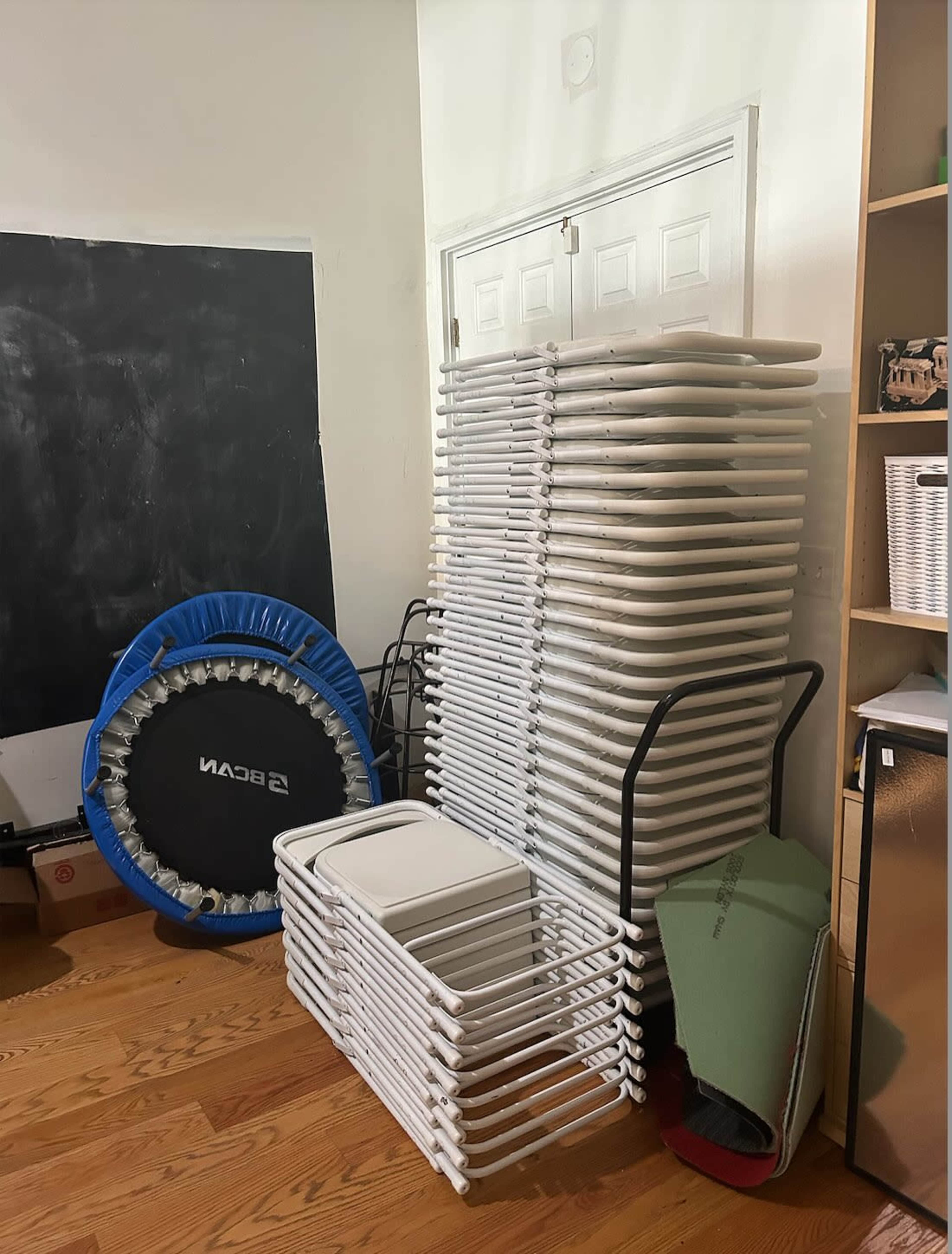The image shows a stack of white folding chairs beside a small trampoline and several other items in a room with wooden flooring.