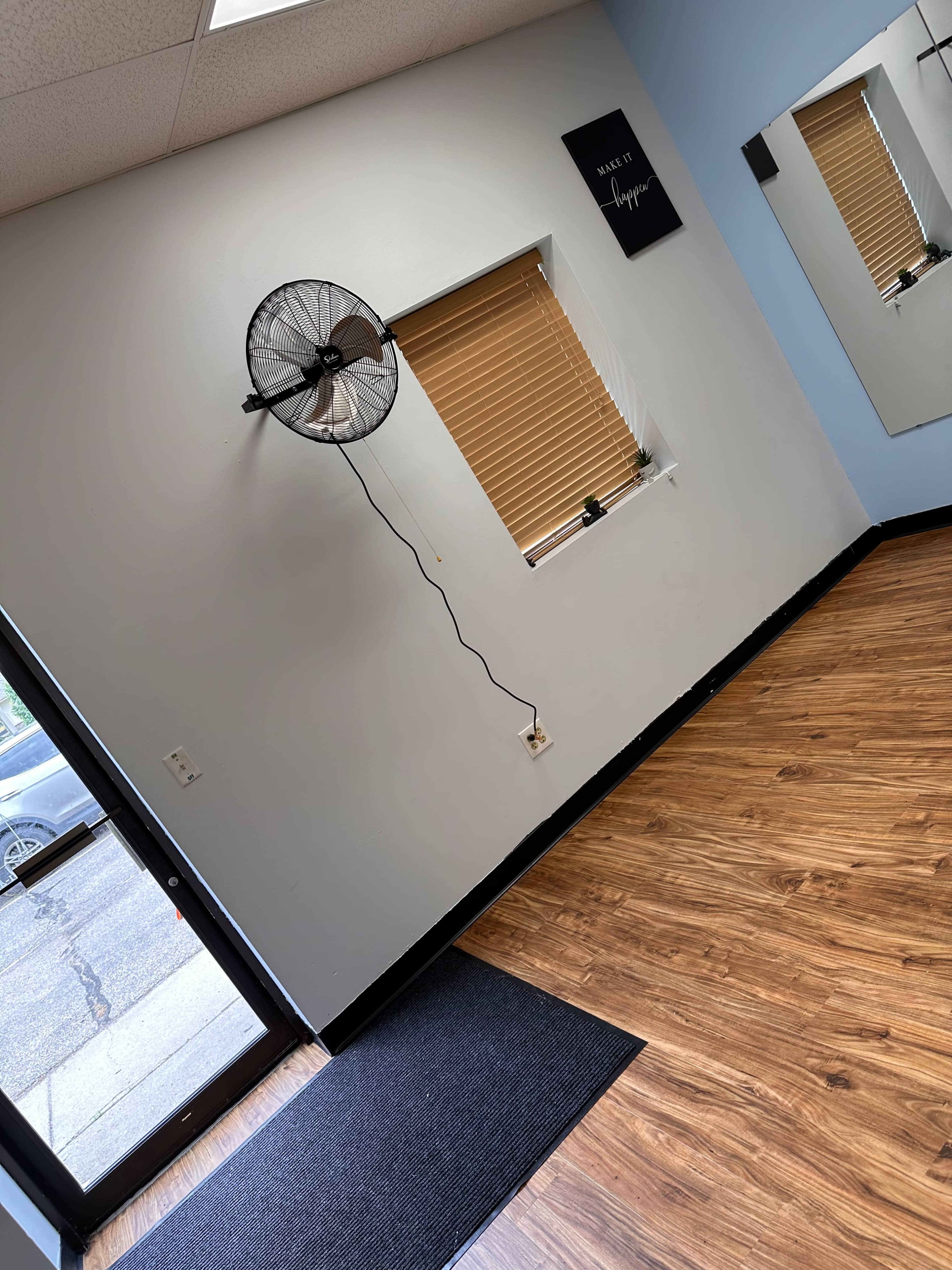 The image shows an empty room with a wall-mounted fan next to a window, a mirror, and a black mat by the entrance.