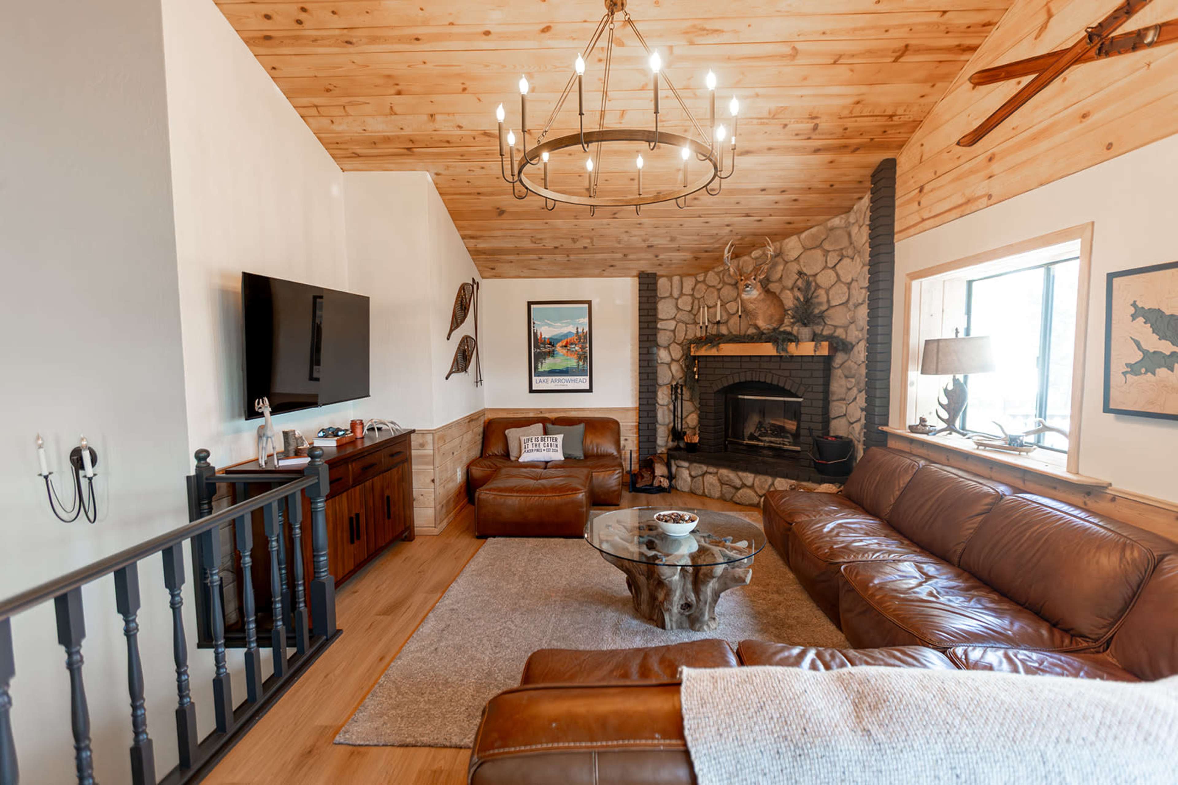 The image shows a cozy living room with wood-paneled walls, a stone fireplace, a large leather sofa, and a chandelier hanging from a wooden ceiling.