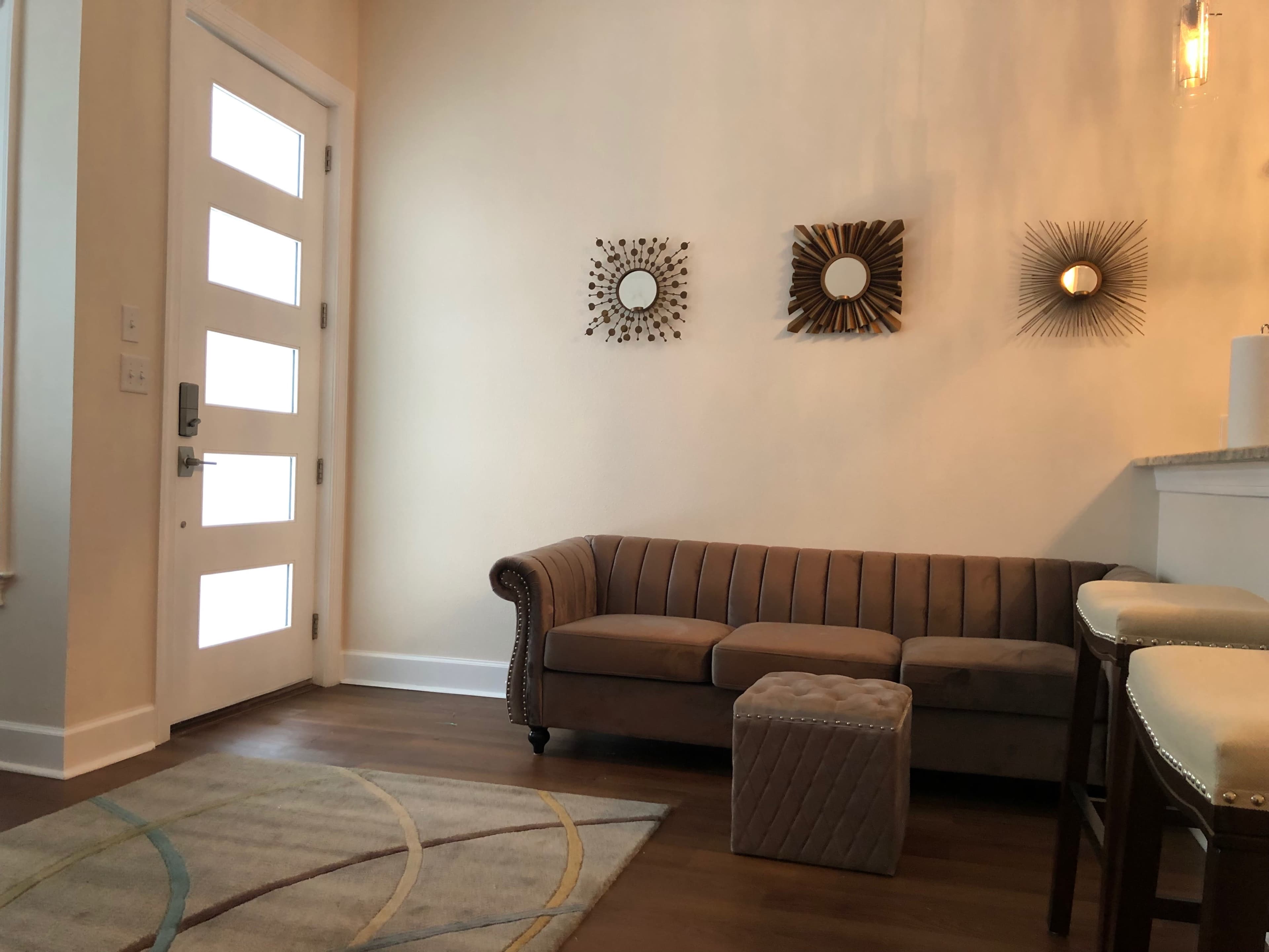A modern entryway featuring a gray tufted couch, a small ottoman, and three decorative wall mirrors above the couch.