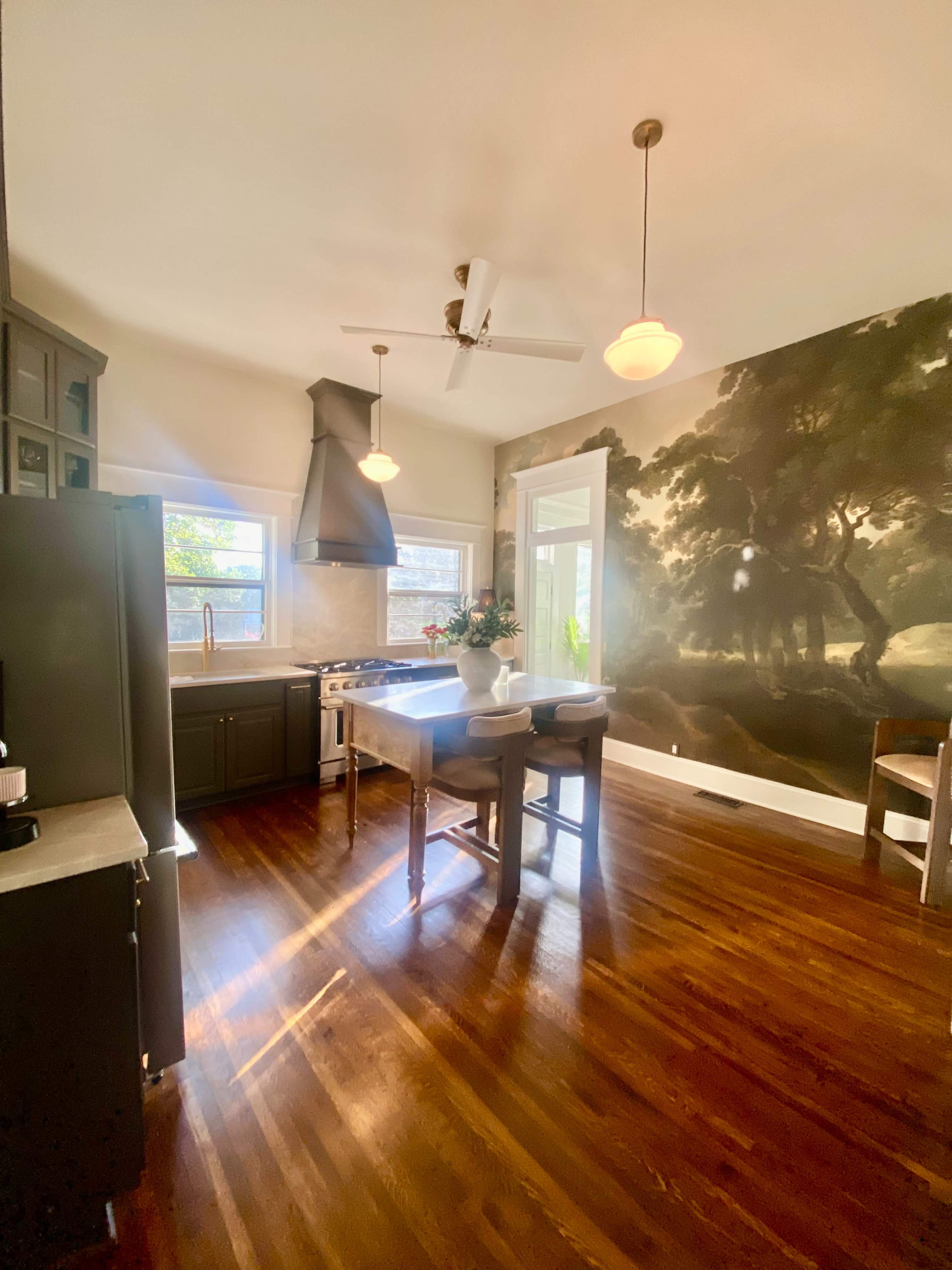 The kitchen features a central island with seating, modern appliances, and a large mural of trees on one wall, illuminated by natural light from the windows.