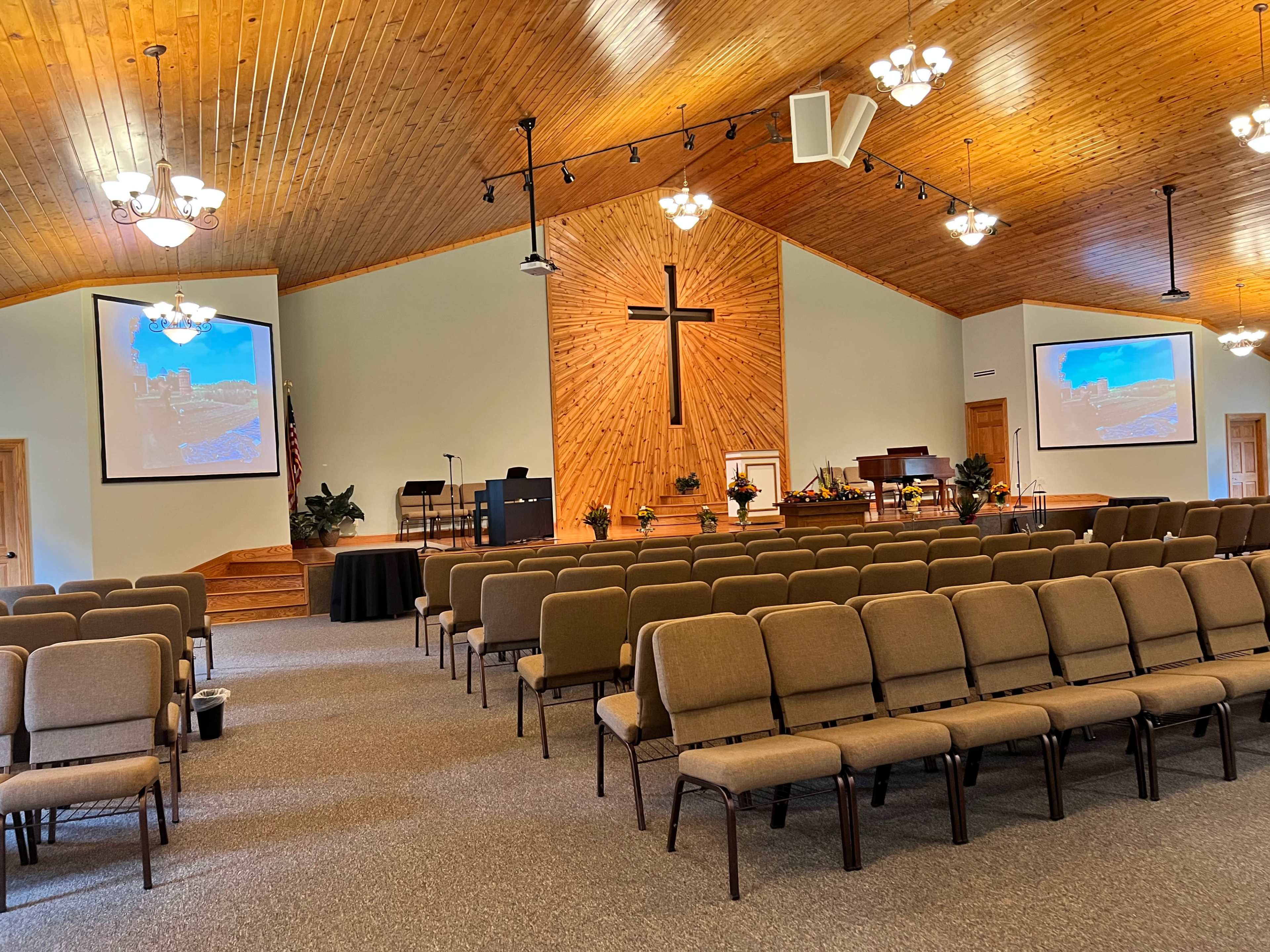 The interior of a church features rows of seated chairs facing a front stage with a large wooden cross, flanked by two projection screens.
