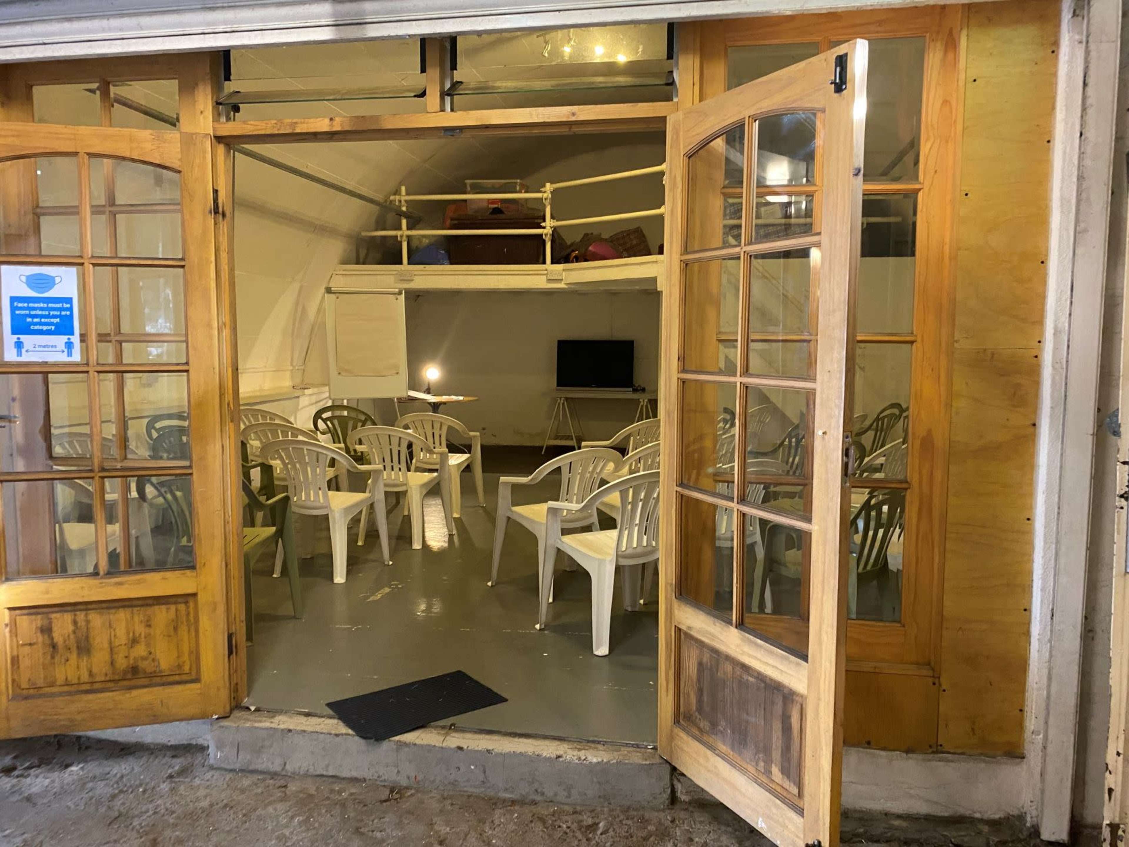 A room with white walls and a wooden floor contains circular tables surrounded by plastic chairs, with a television on one side and doors open to the outdoor area.