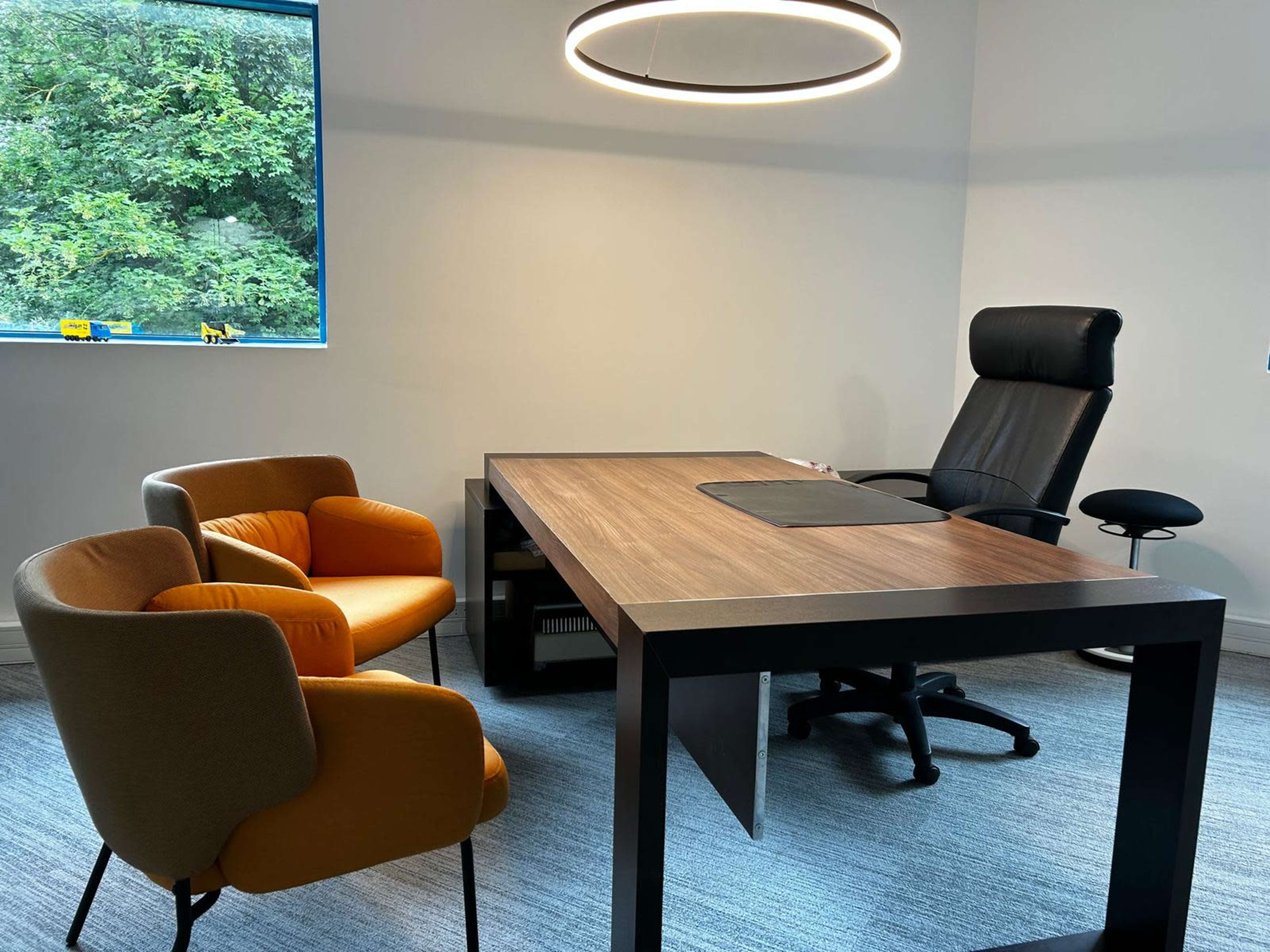 The image shows a modern office interior with a large wooden desk, a black swivel chair, and two accent chairs in orange and beige, situated near a window with greenery visible outside.