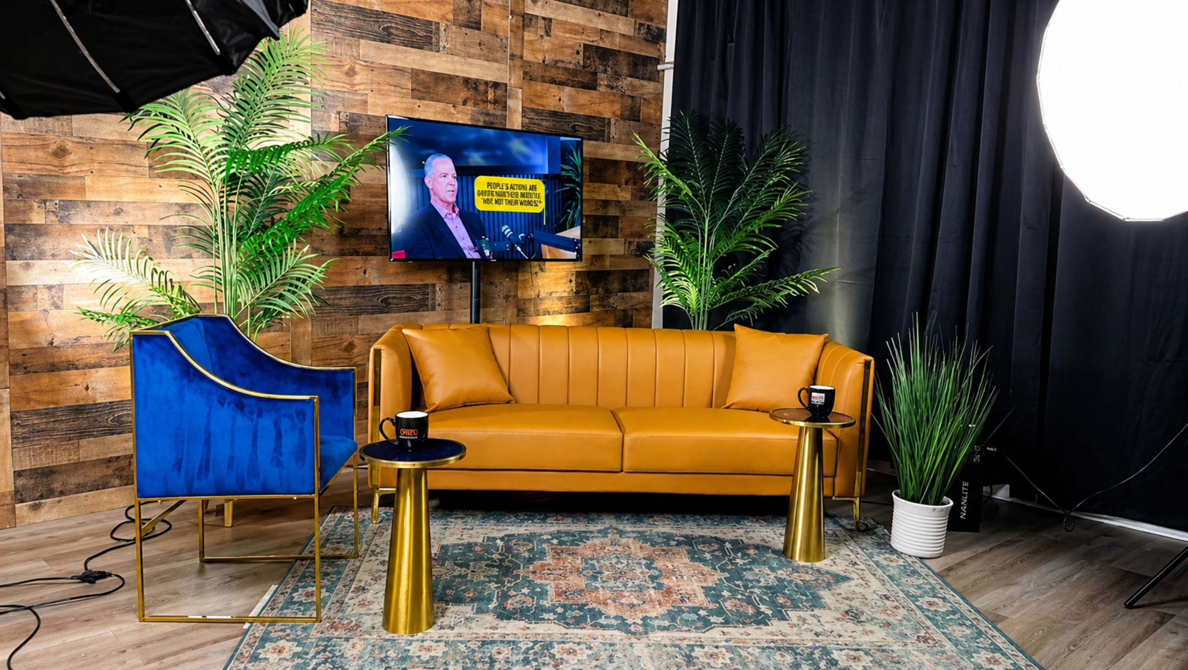 The image shows a styled interior set featuring a mustard-colored sofa, a blue accent chair, two gold side tables, and potted plants, with a TV displaying a news presentation in the background.