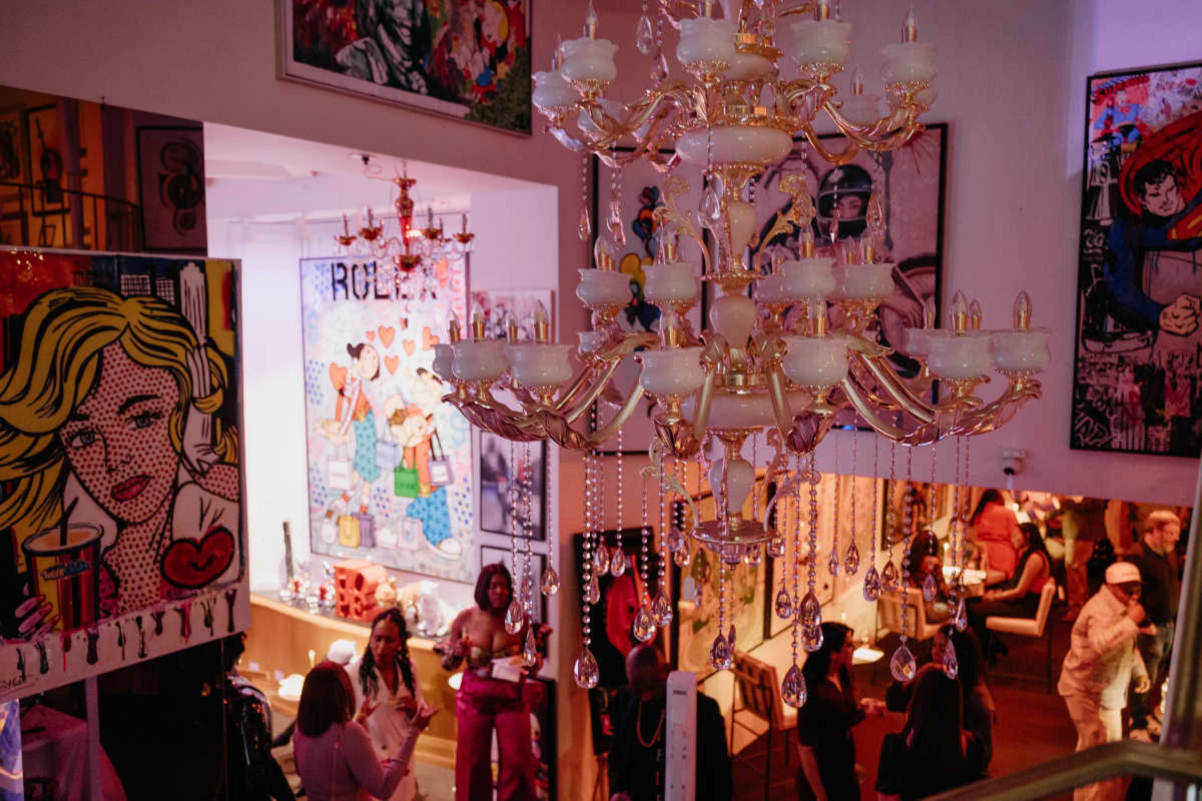The image shows a vibrant art gallery filled with colorful artwork, a large chandelier hanging from the ceiling, and visitors engaging with the exhibits.
