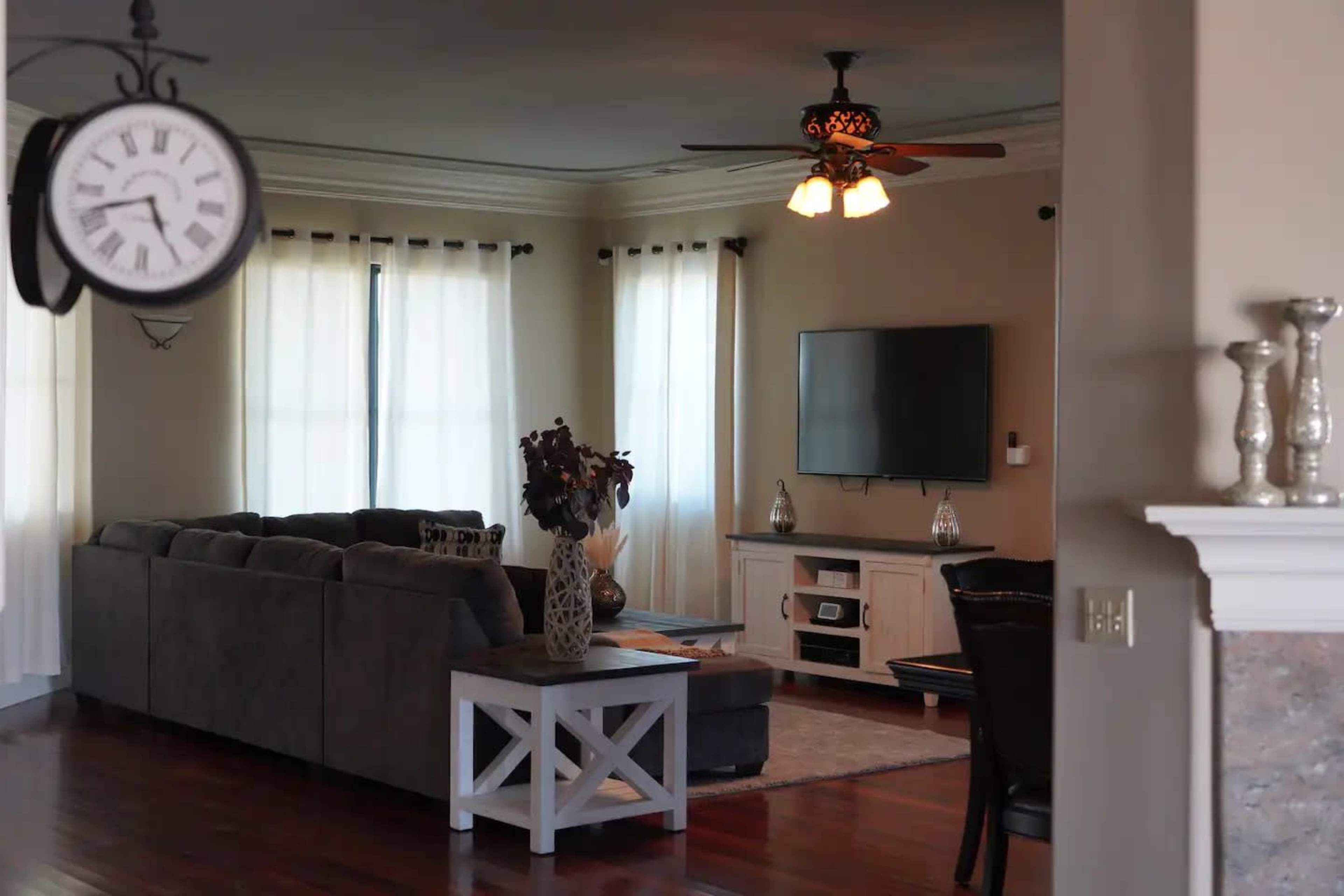 A living room with a gray sectional sofa, a coffee table, and a mounted television on the wall beneath two large windows with sheer curtains.
