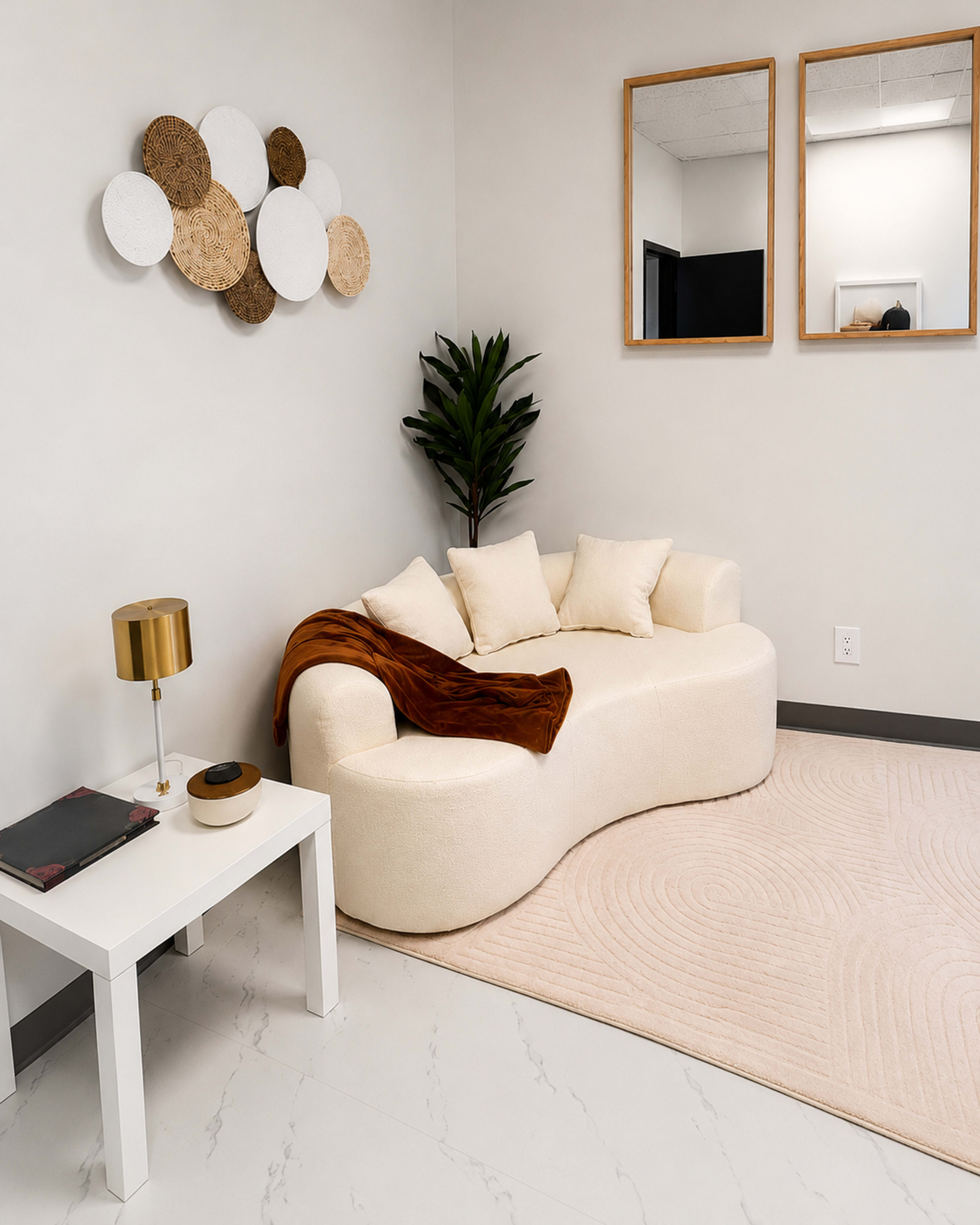 The image shows a modern living space featuring a curvy beige sofa, a small white side table with a lamp, and decorative wall art made of circular materials.