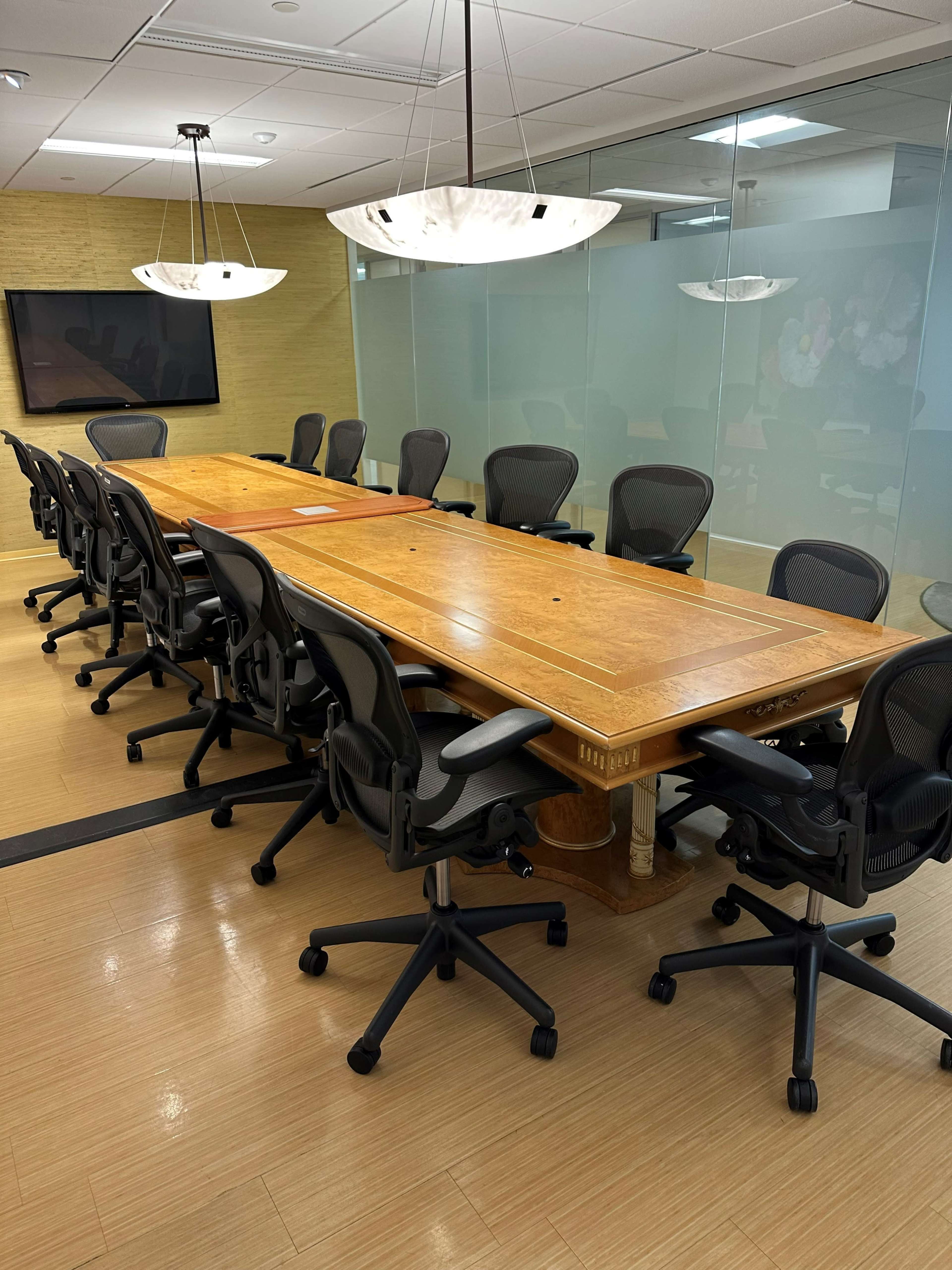 A large wooden conference table is surrounded by several office chairs in a modern meeting room with glass walls.