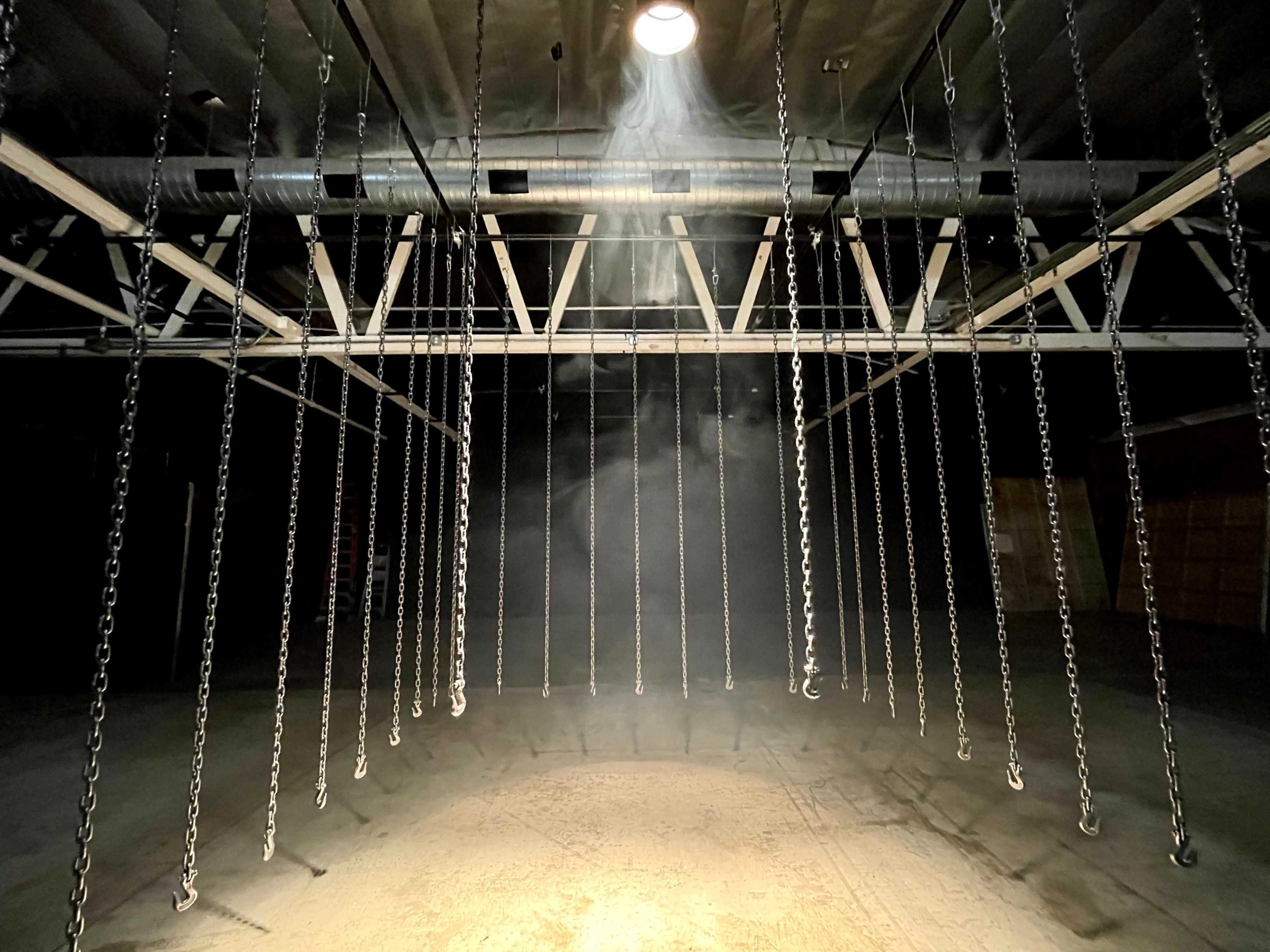 A dimly lit industrial space with metal chains hanging from a ceiling beam, illuminated by a single overhead light creating a beam of light through the surrounding dust.