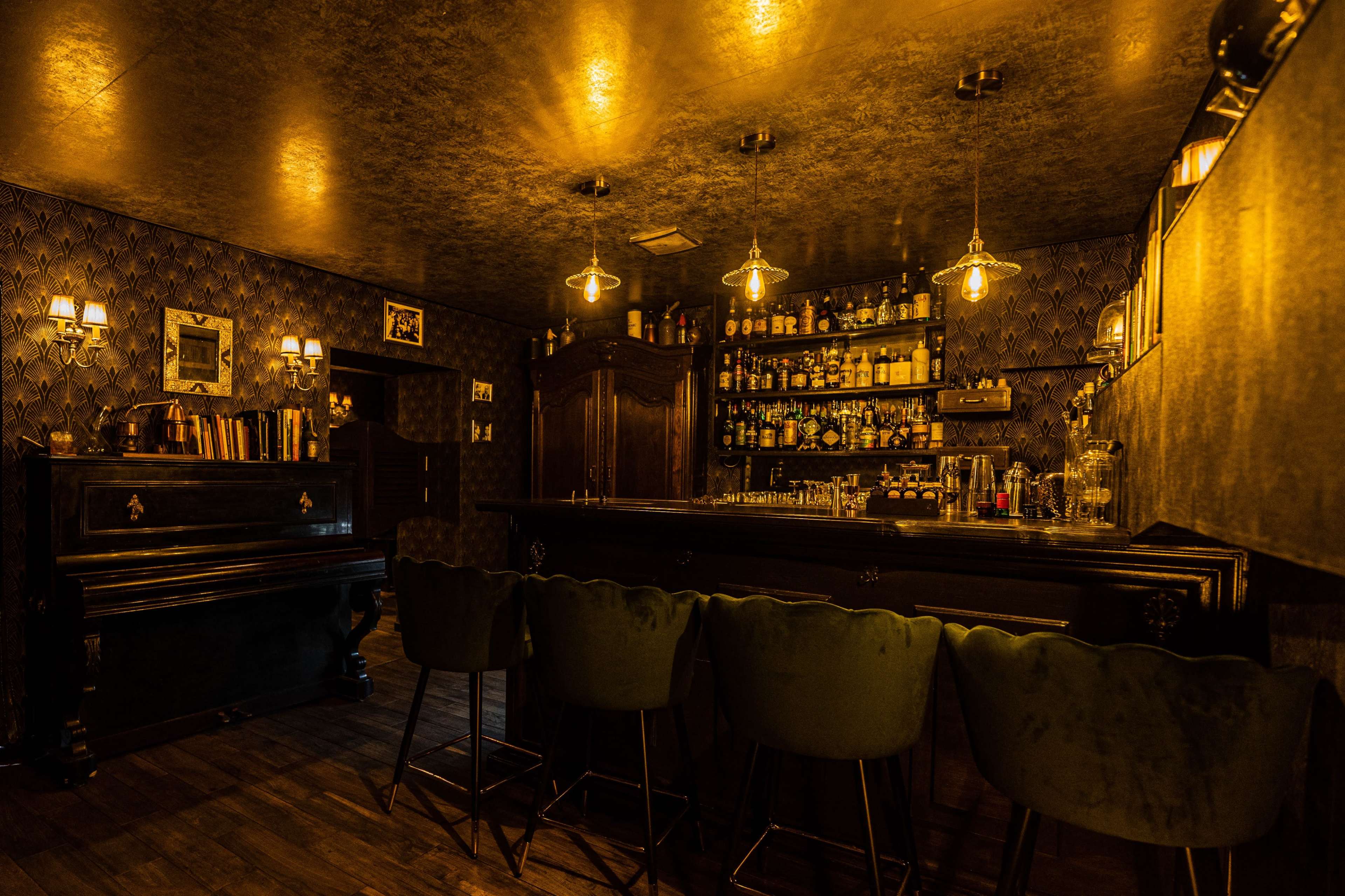 The image shows a dimly lit bar with a dark wooden counter, green upholstered bar stools, a piano in the corner, and a shelf filled with various bottles and glasses.