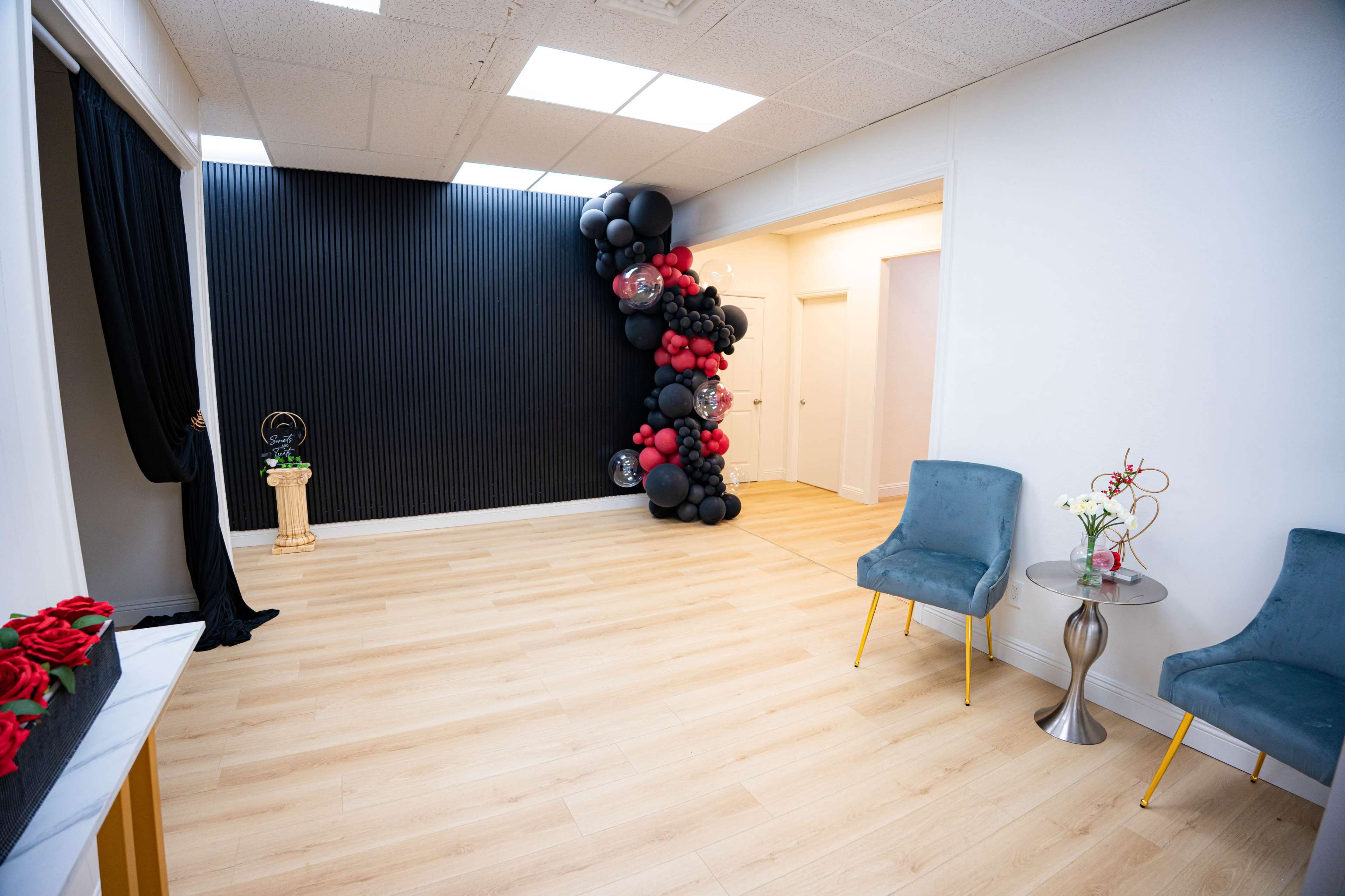 The image shows a modern, empty room featuring light wooden flooring and a black accent wall adorned with a cluster of red and black balloons, along with two blue chairs and a small table with a vase of flowers.