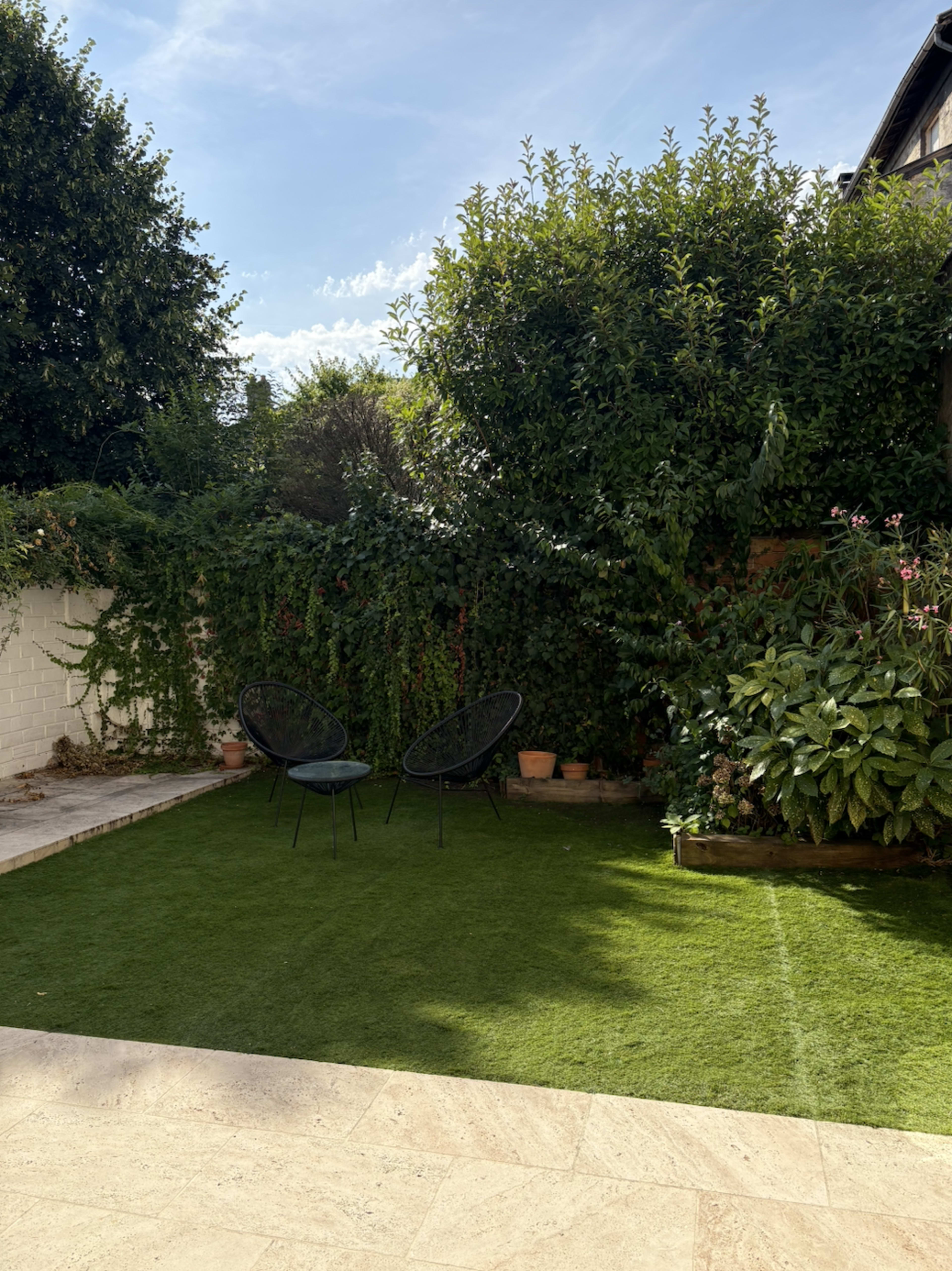 The image shows a garden area with two black chairs on artificial grass, surrounded by lush greenery and a brick wall.