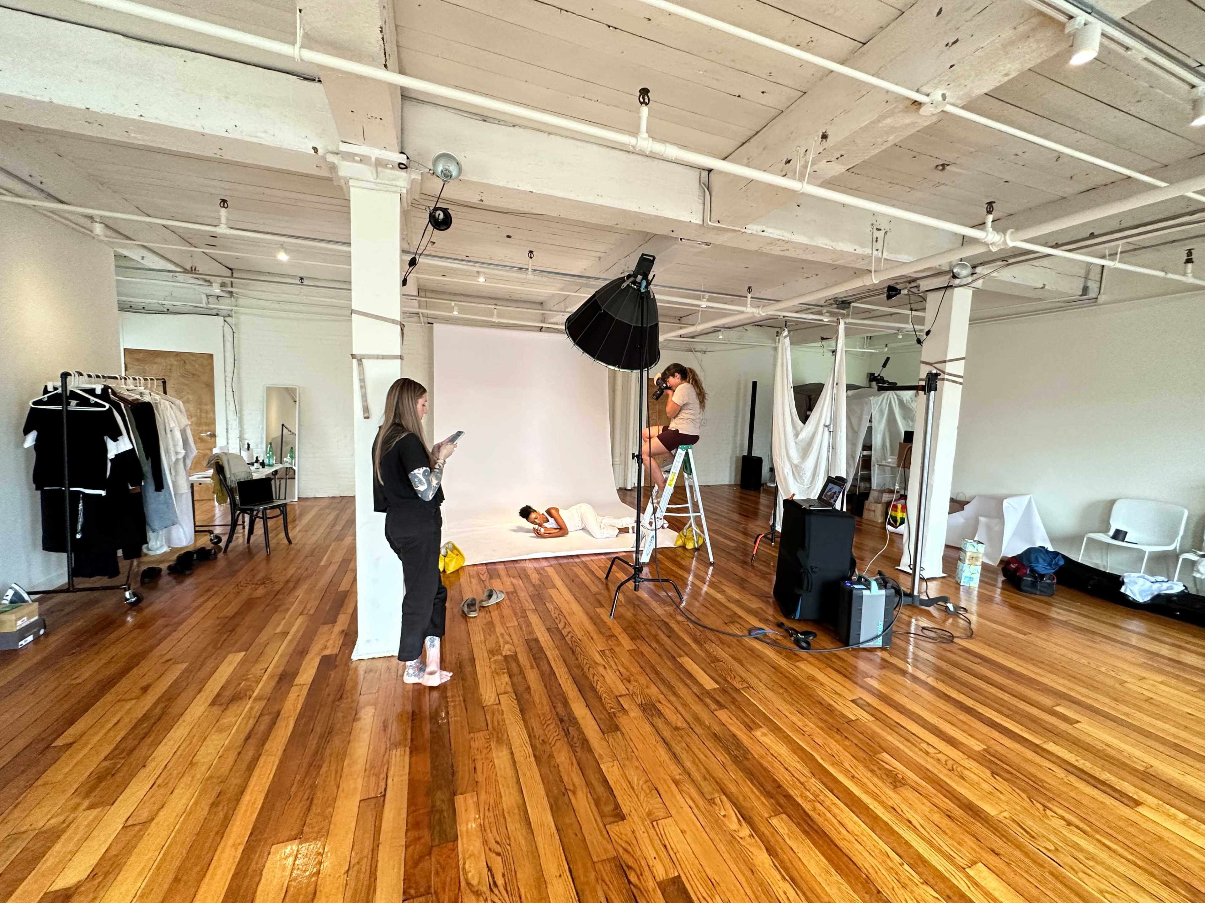 A photography studio features a model posing on a backdrop while a photographer adjusts lighting equipment, with staff organizing clothing and accessories.
