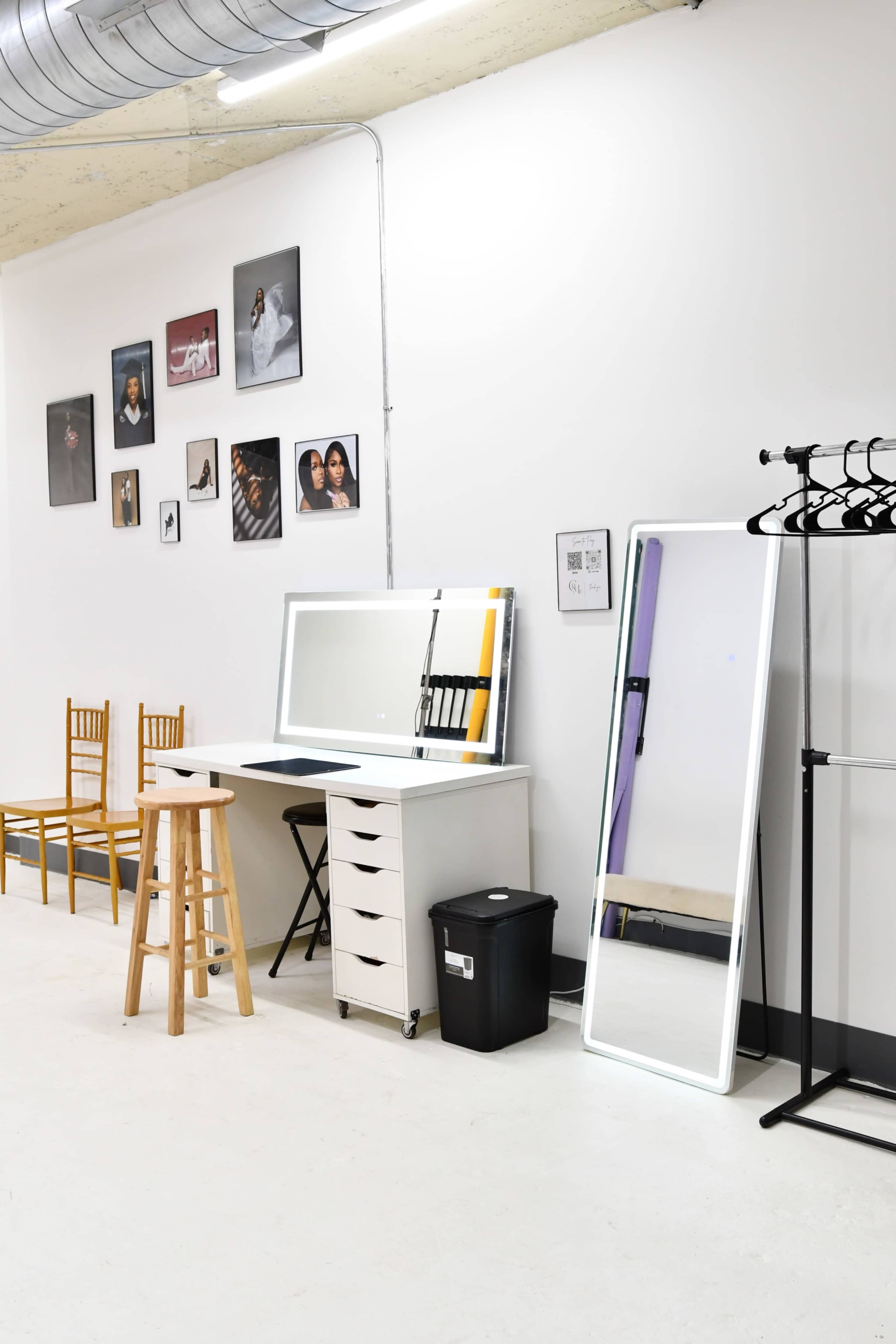 The image shows a minimalist indoor space featuring a vanity table with lights, a large mirror, a stool, and a collection of framed photographs on the wall.
