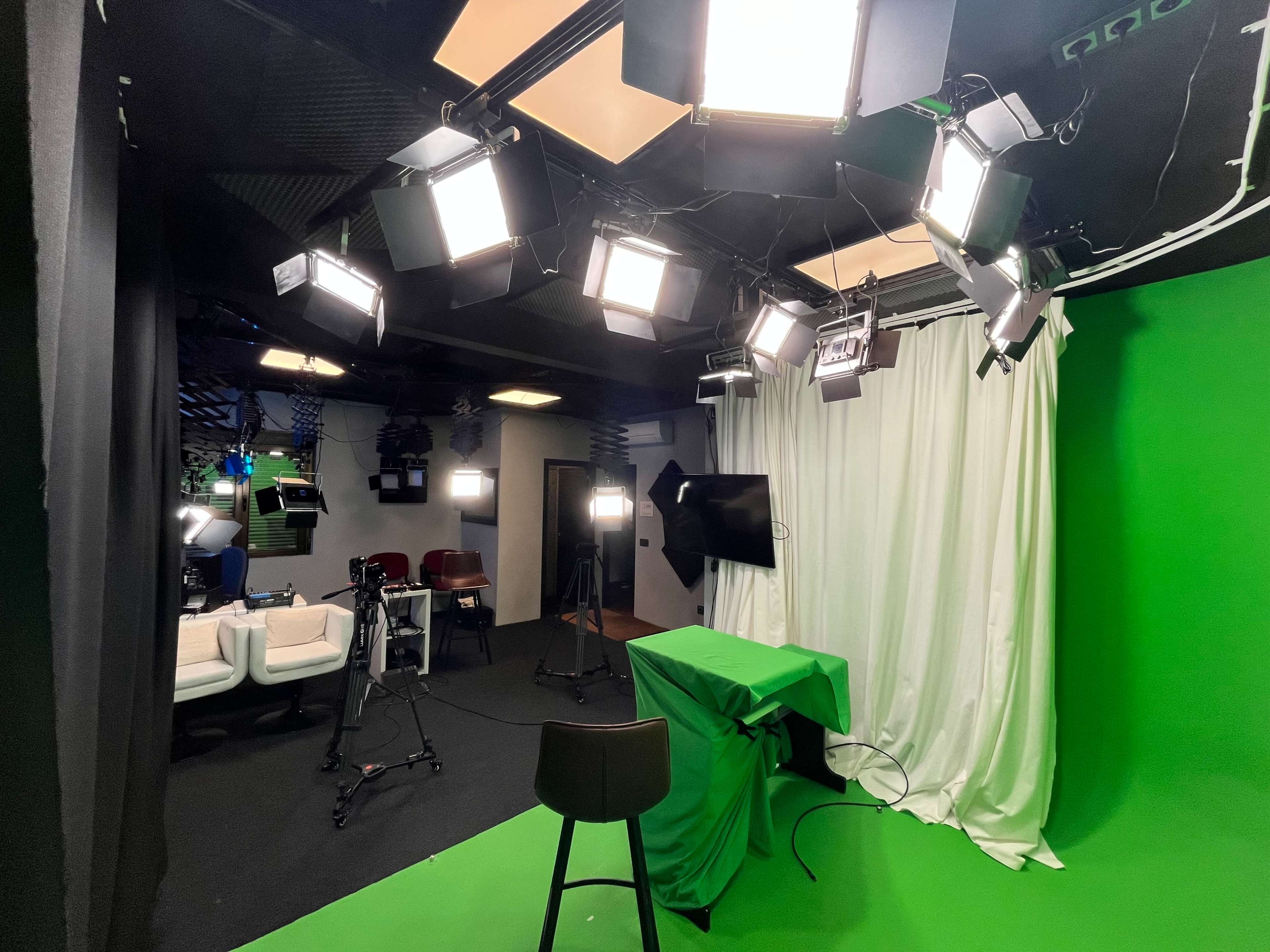 The image shows a video recording studio with multiple lights, a green backdrop, and a chair in front of an empty table.
