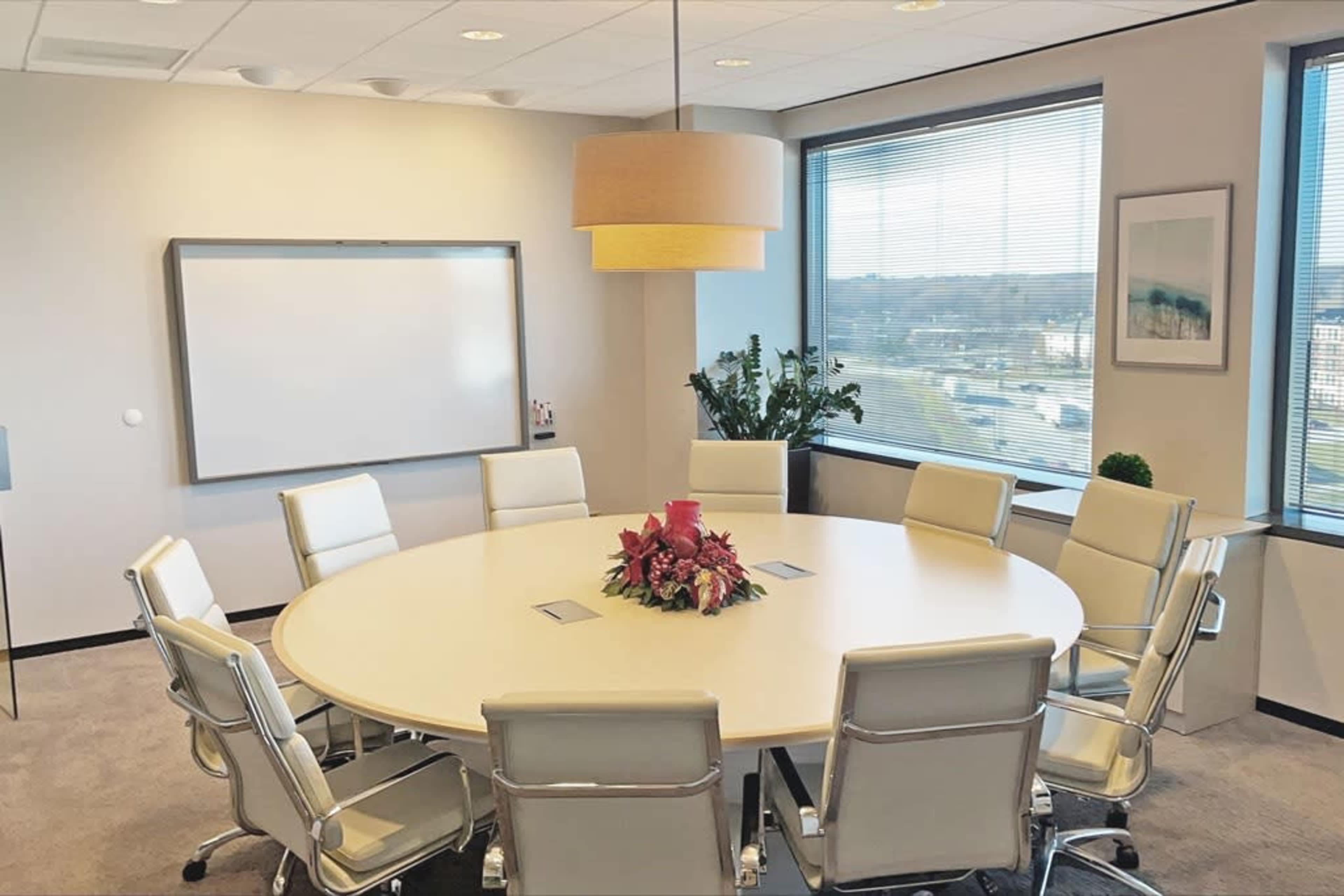 A round conference table surrounded by white chairs is set up in a well-lit meeting room with large windows.