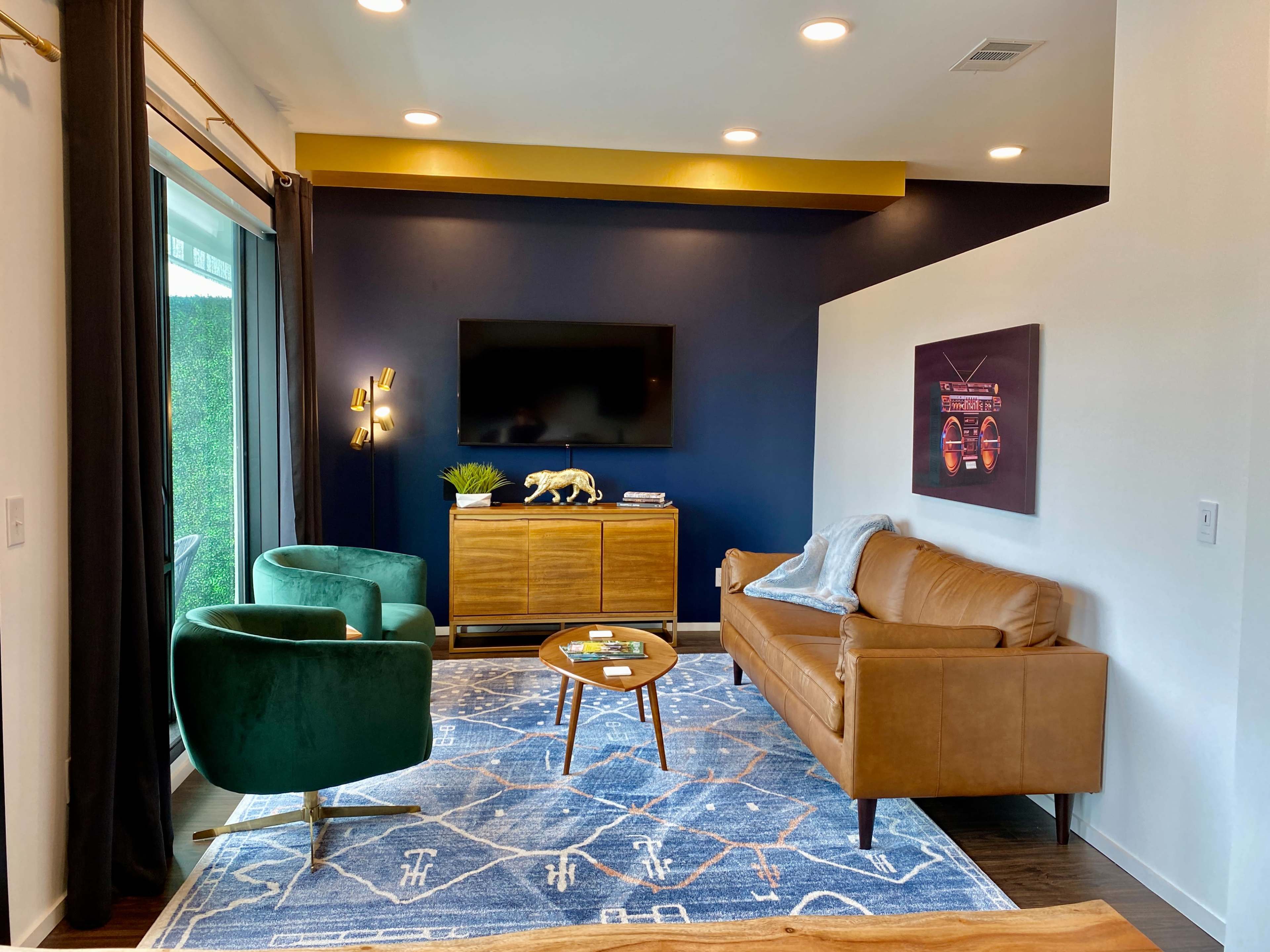 A modern living room featuring a brown leather sofa, green chairs, a wooden coffee table, and a large television mounted on a dark blue wall.