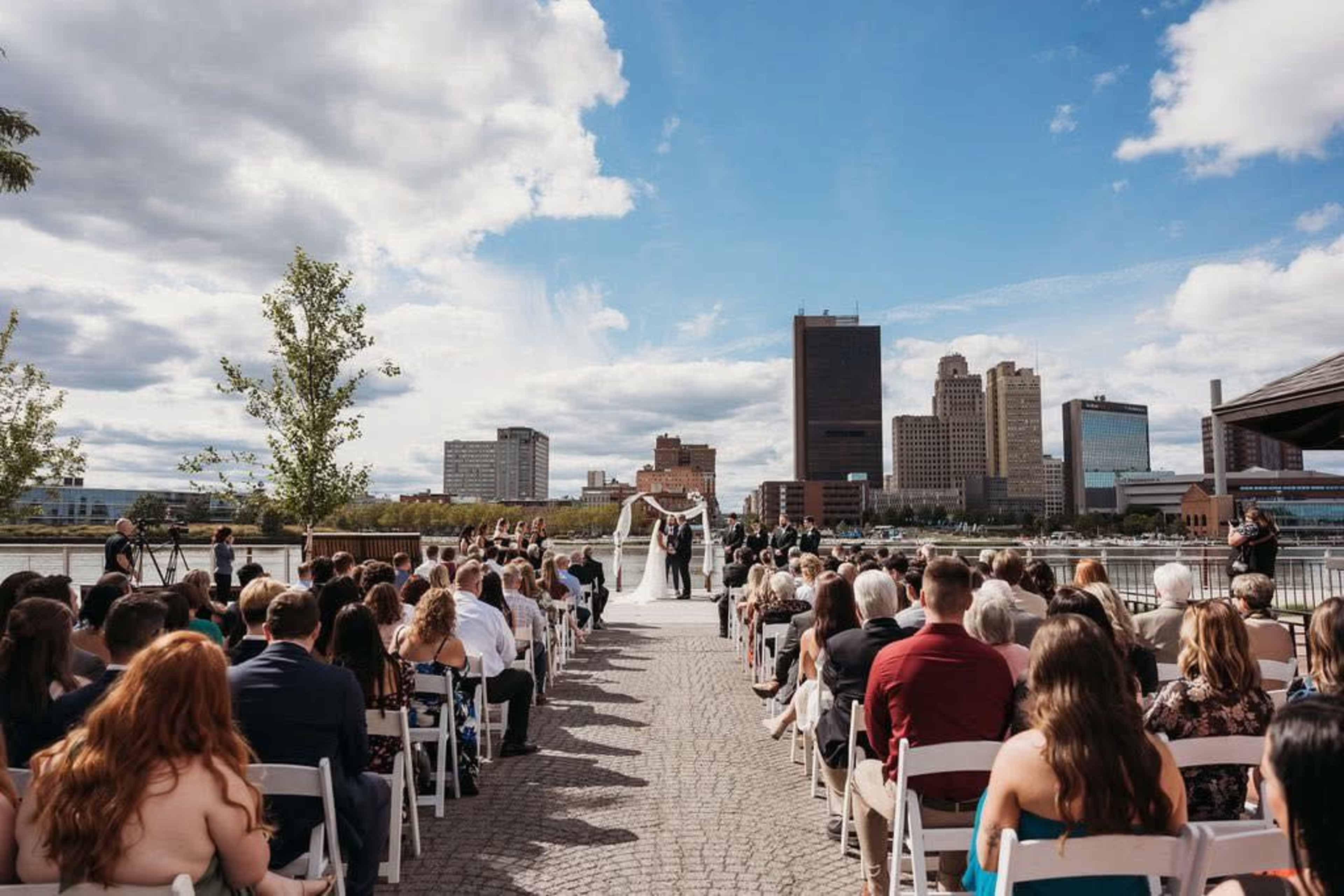 A wedding ceremony is taking place outdoors with guests seated in chairs, facing the couple standing under an arch, set against a city skyline.