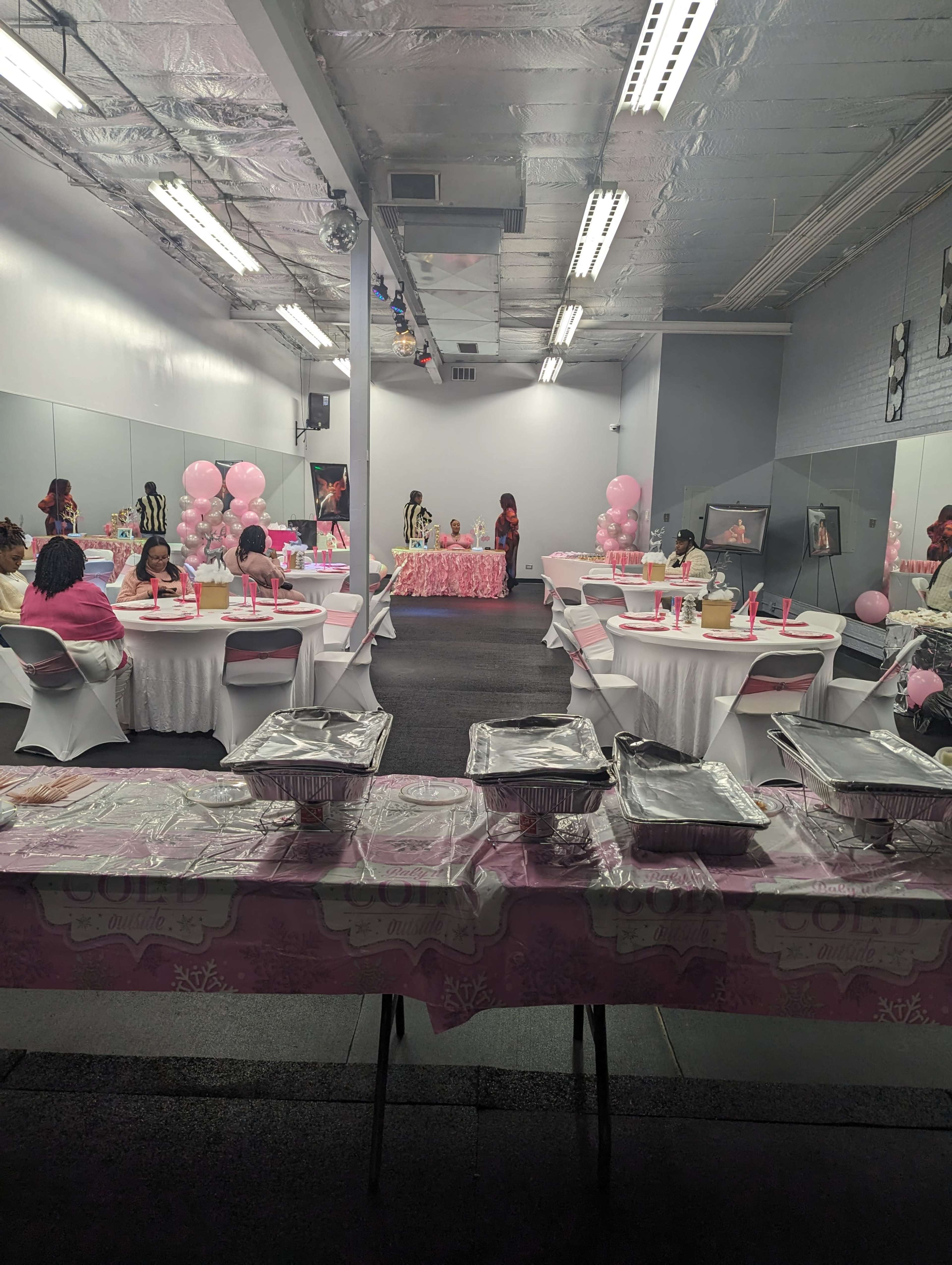 A room is set up for a celebration, featuring tables covered in pink tablecloths, covered dishes, and pink balloons arranged decoratively.