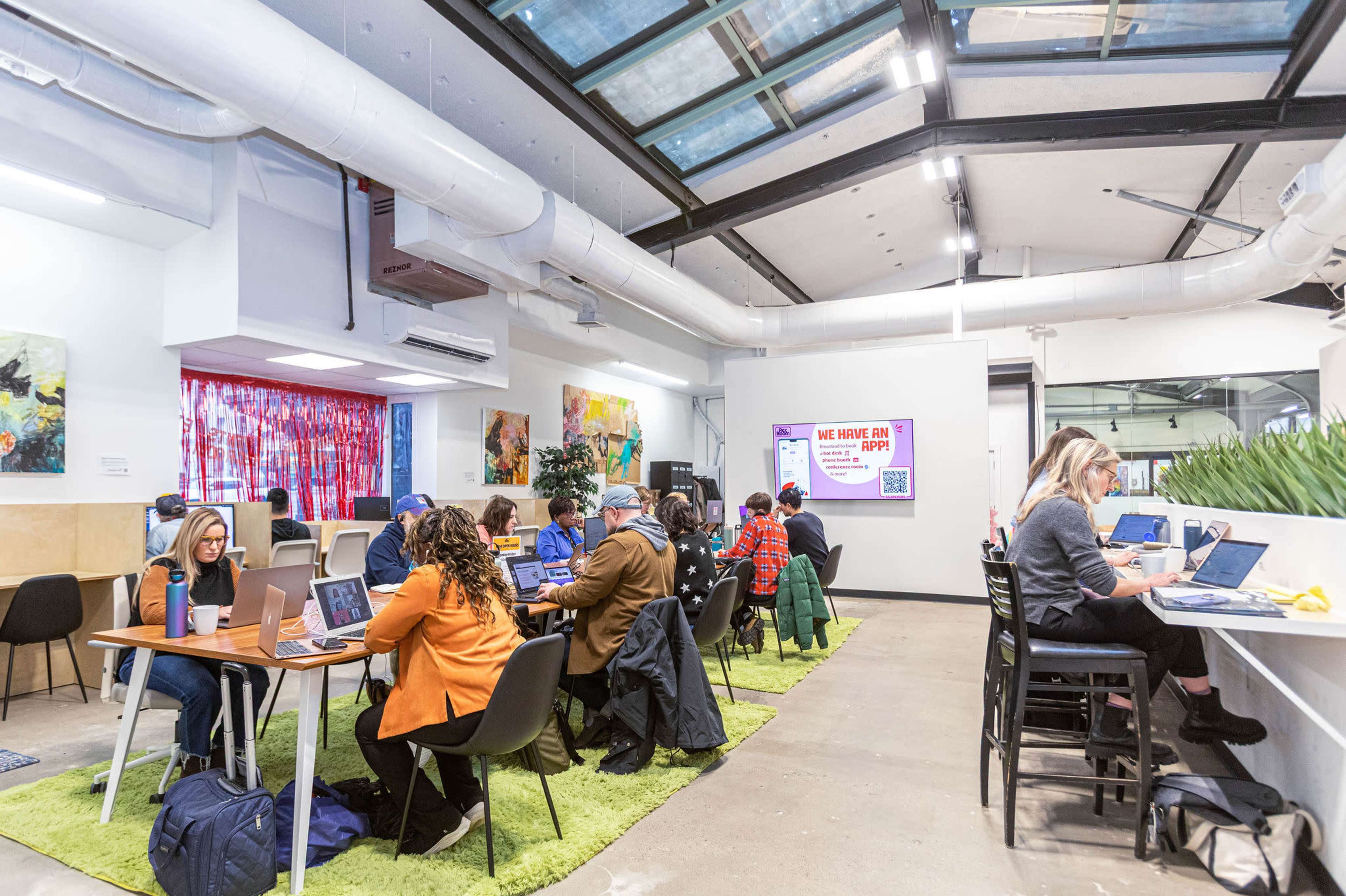 The image shows a modern coworking space with several people working at tables and using laptops, surrounded by vibrant artwork and plants.