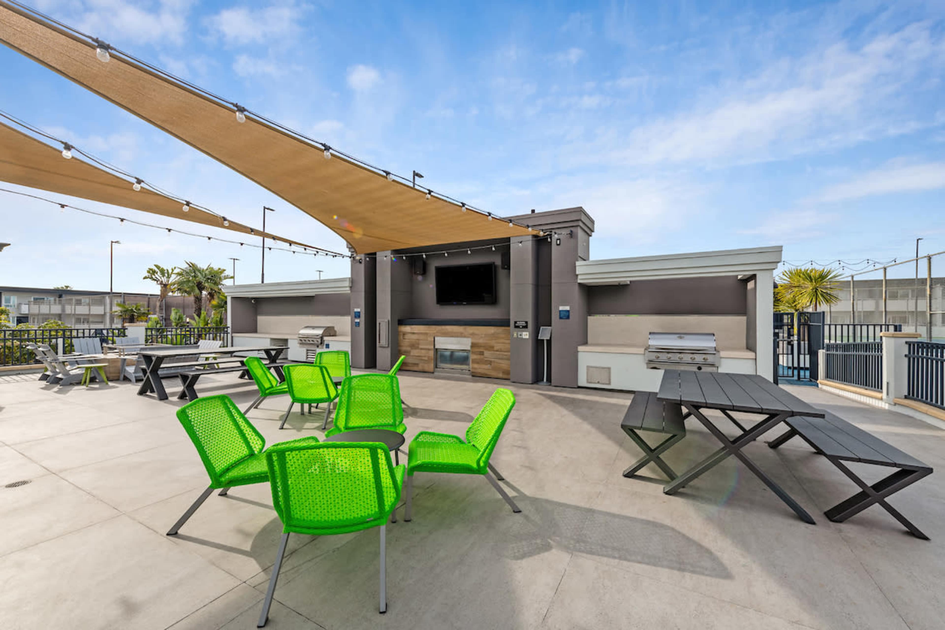 A modern outdoor patio area with green chairs, picnic tables, a grill, and a large screen for viewing.