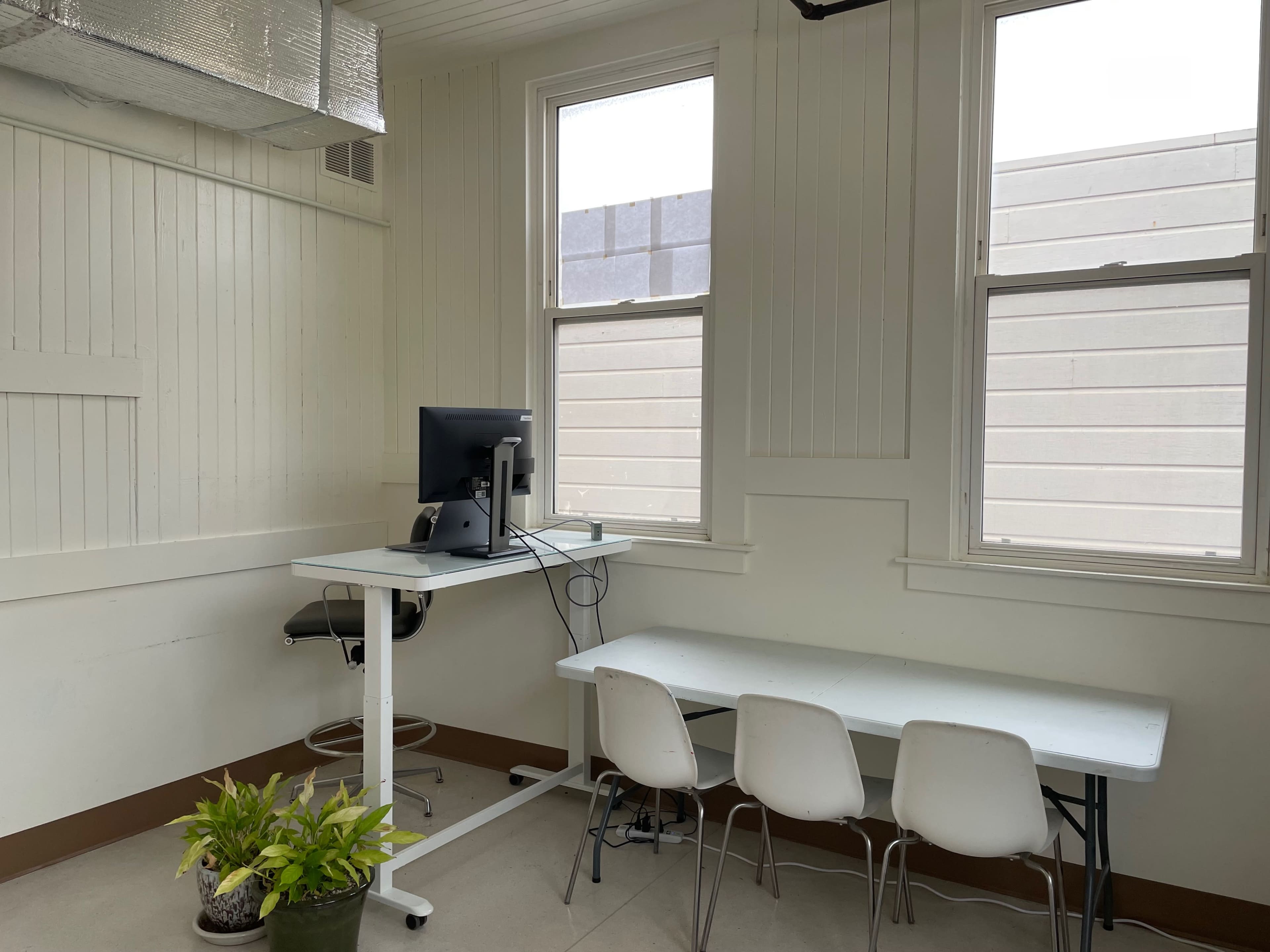 A minimalist room with a computer on a desk, a stool, a table, and three chairs, along with a potted plant.