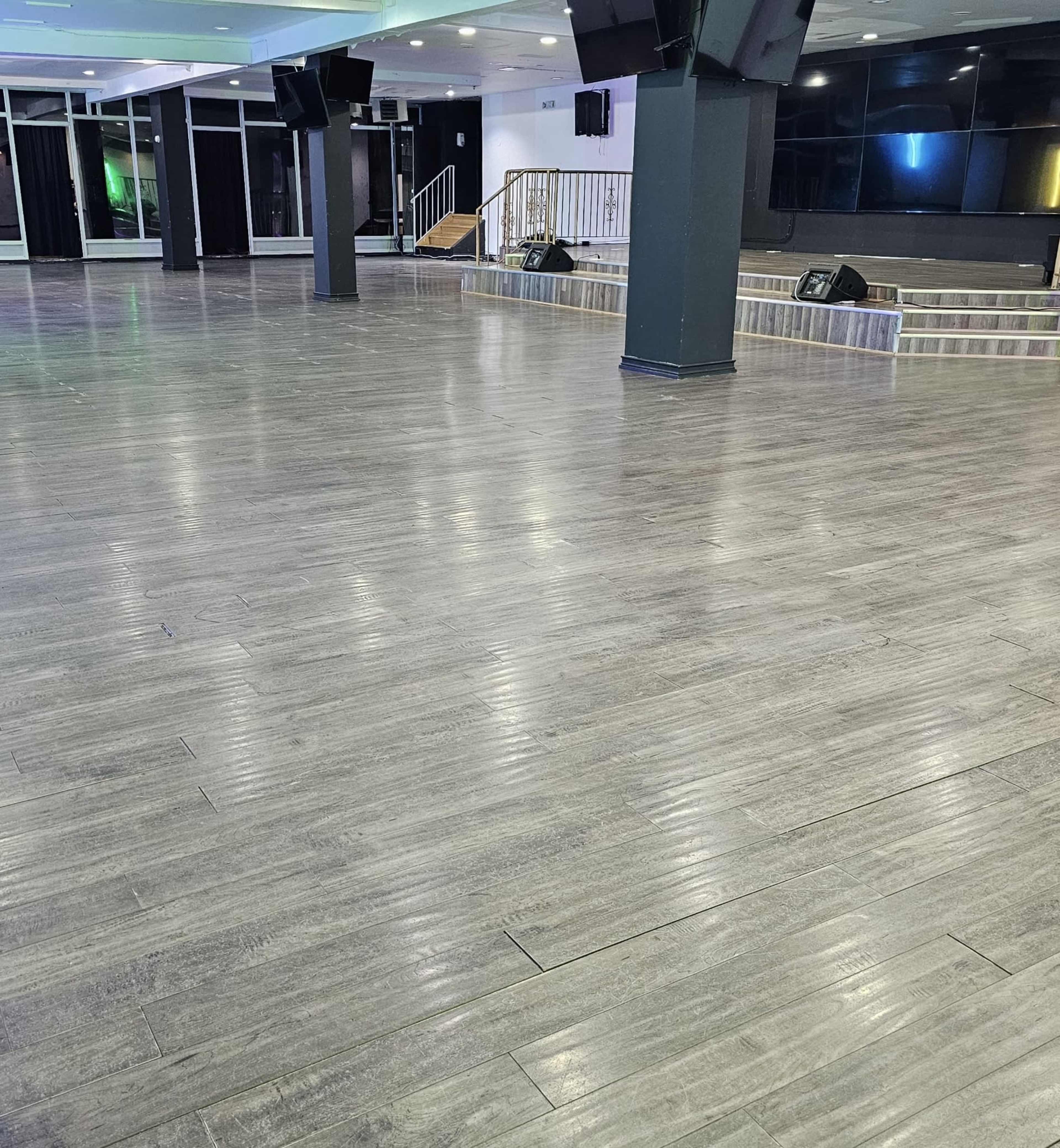 The image shows a spacious indoor venue with a polished wooden floor and a stage area, illuminated by colorful lights.