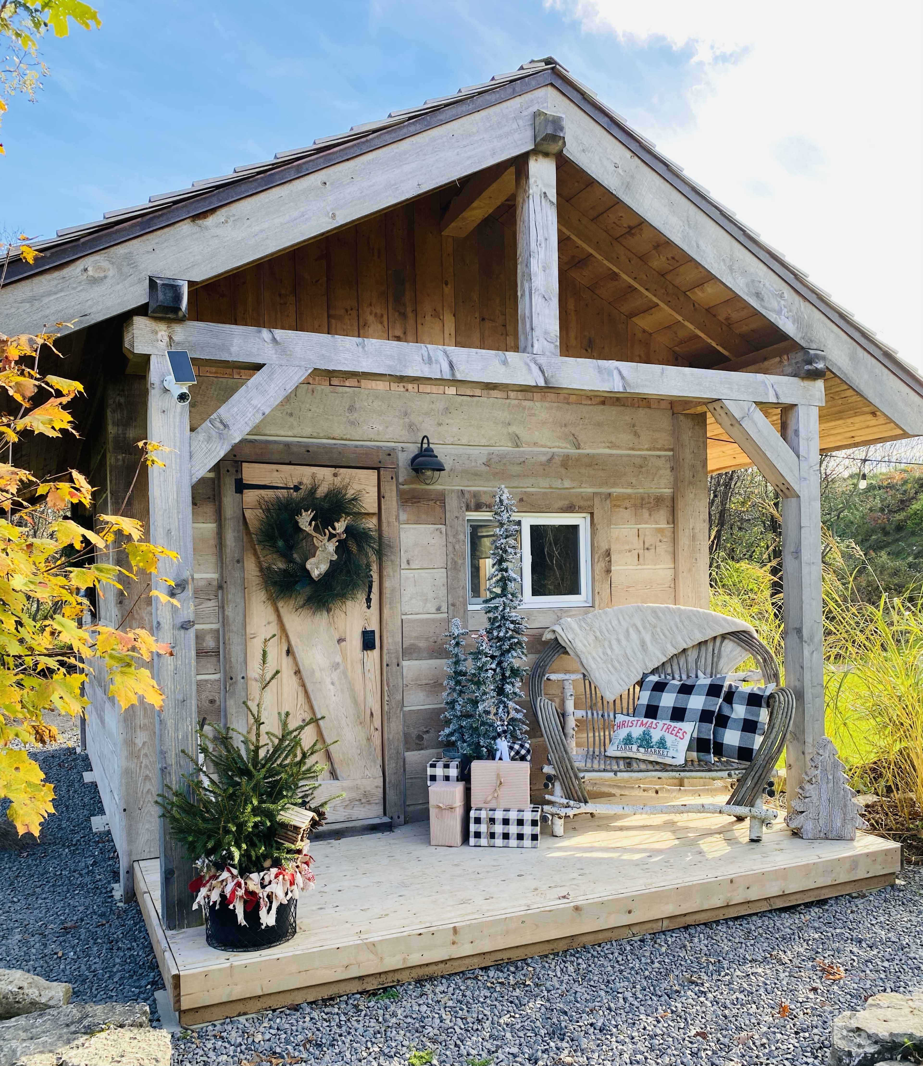 A rustic wooden cabin features a front porch decorated with a wreath, a small Christmas tree, and wrapped presents.