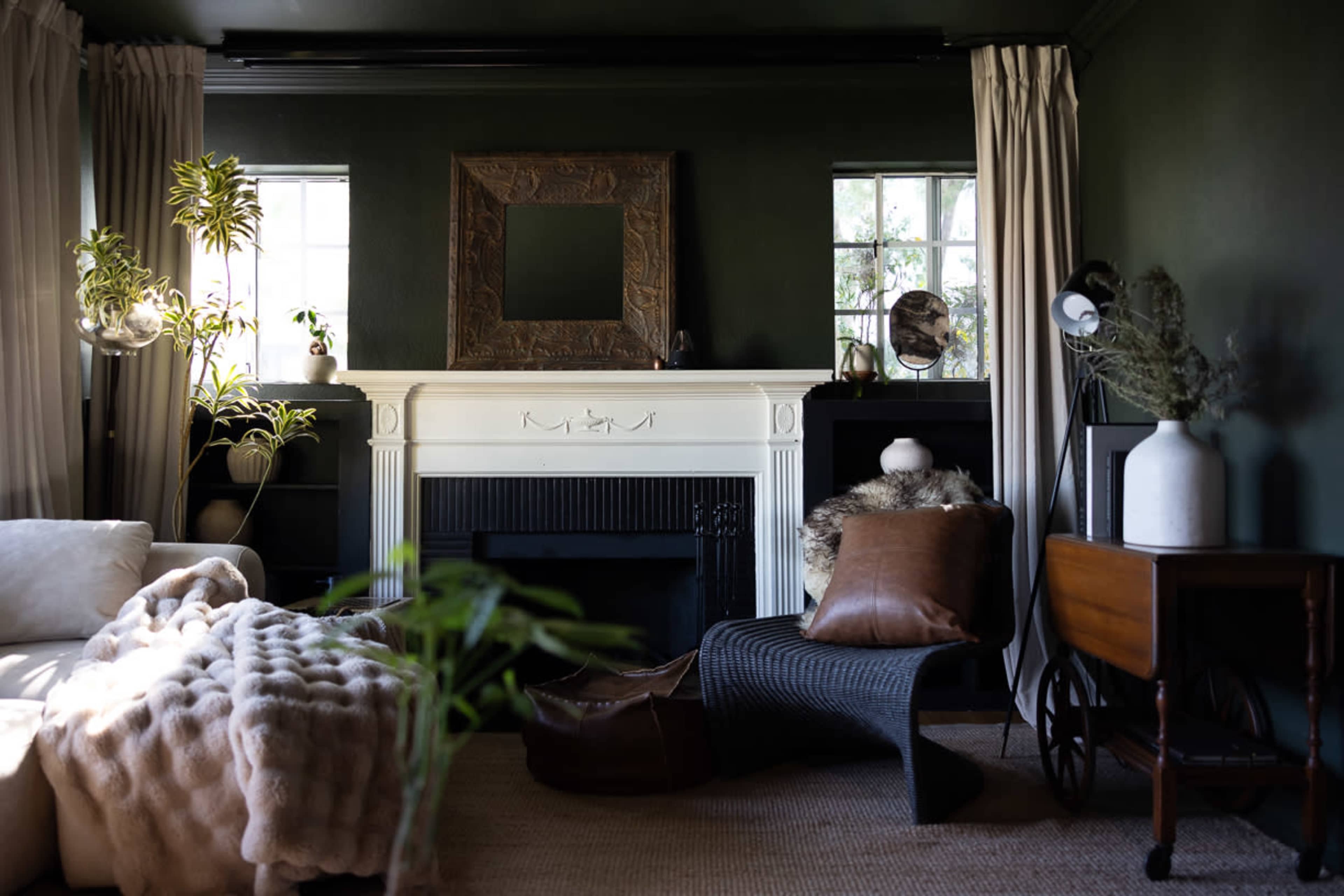 A cozy living room features a white fireplace, dark green walls, a large mirror, and a mix of plants and furniture.