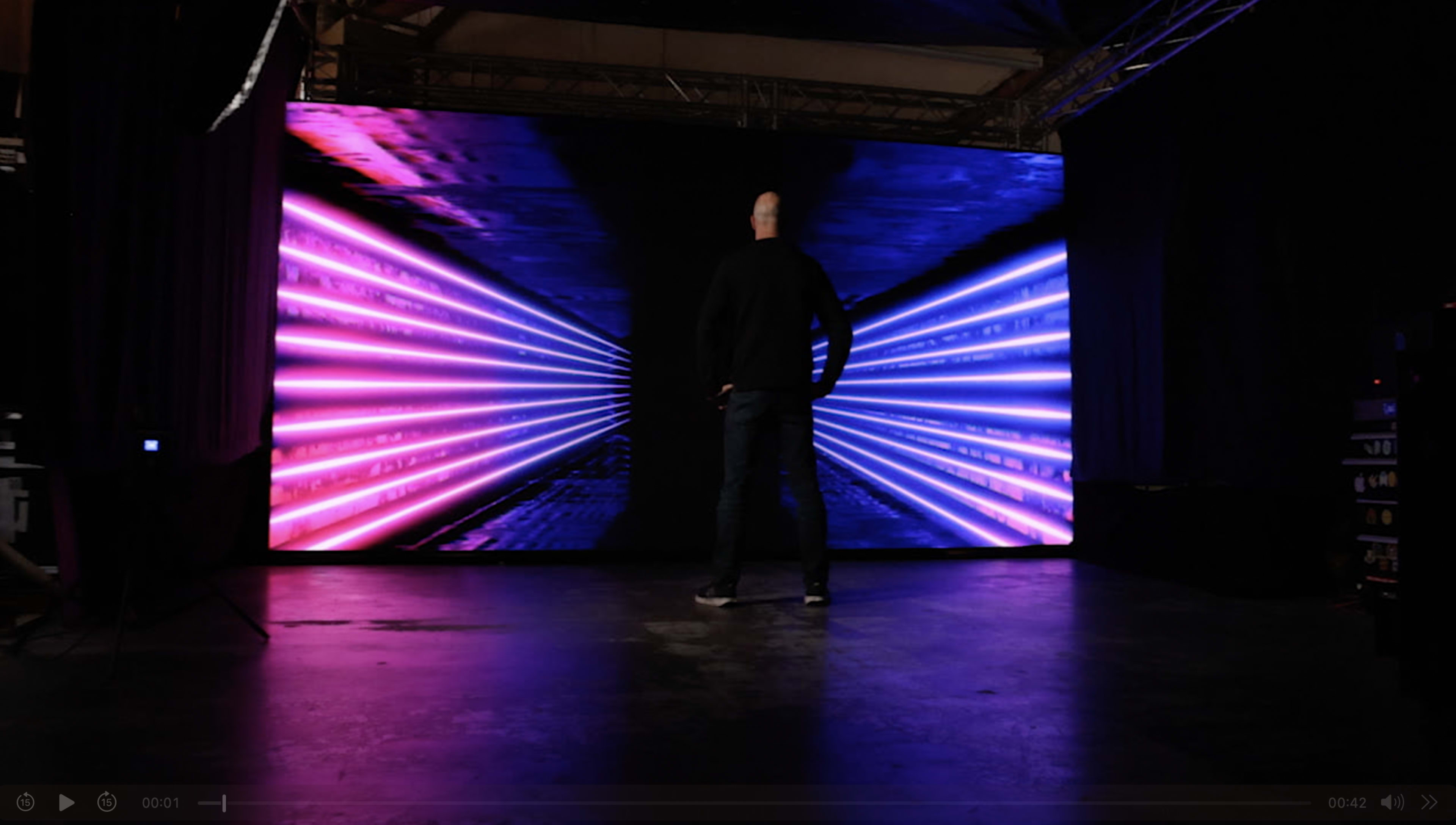 A person stands in front of a large screen displaying vibrant blue and pink light patterns in a dark room.