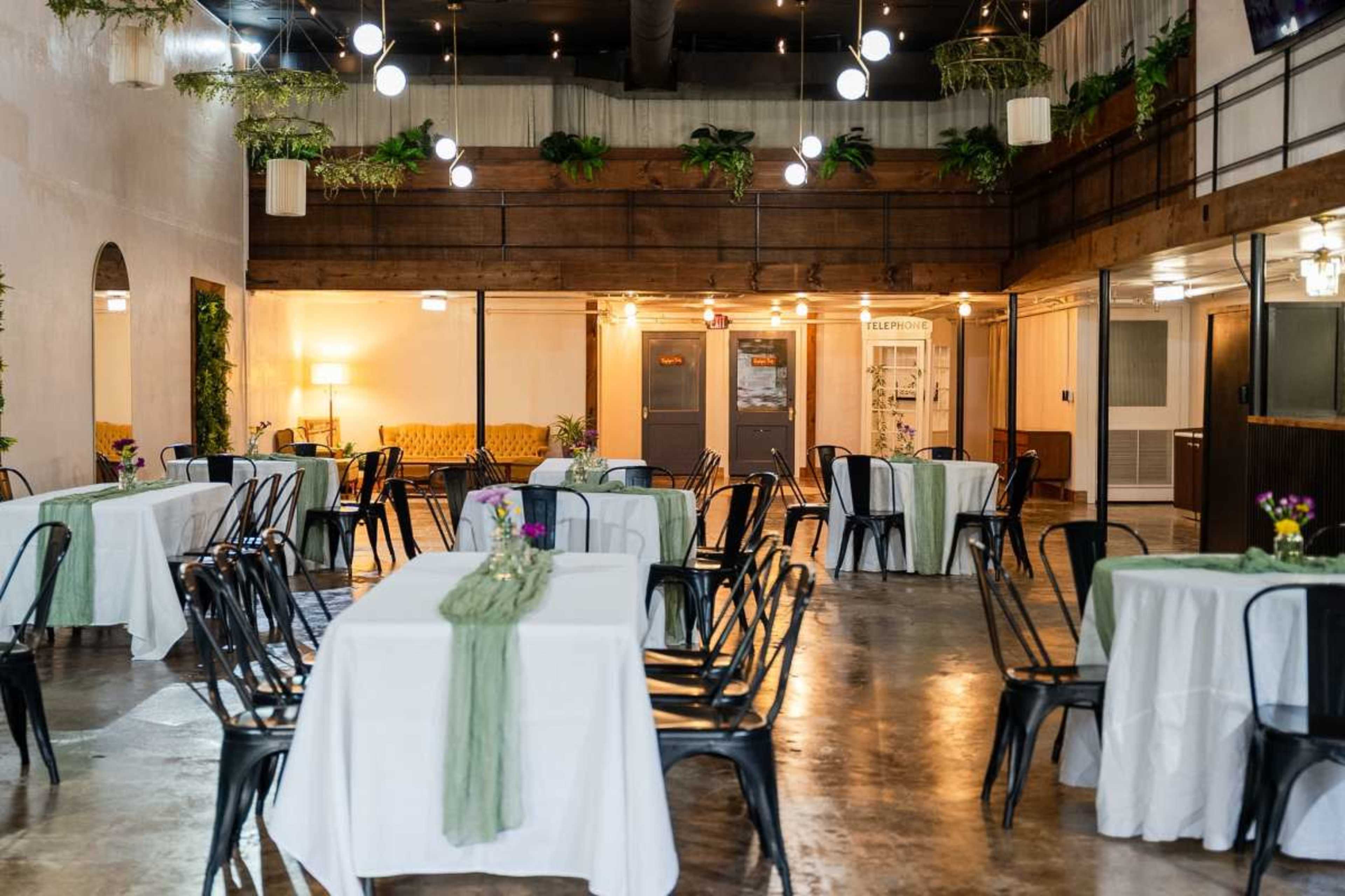 The image shows a spacious event venue with several round tables covered in white tablecloths and adorned with small floral centerpieces, surrounded by wooden beams and greenery.