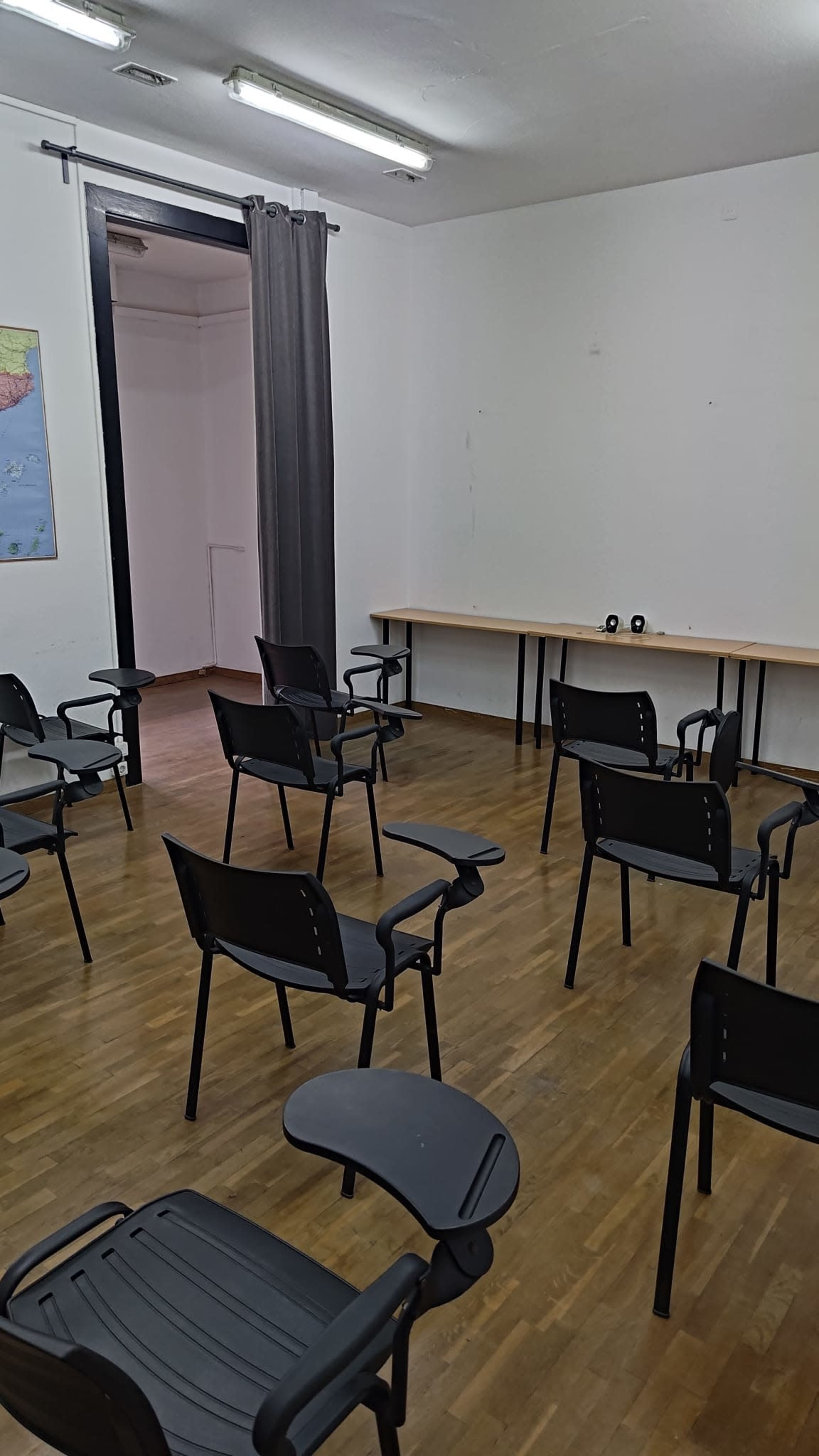 The image shows a room with several black chairs arranged in a semicircle facing a long table against a white wall.