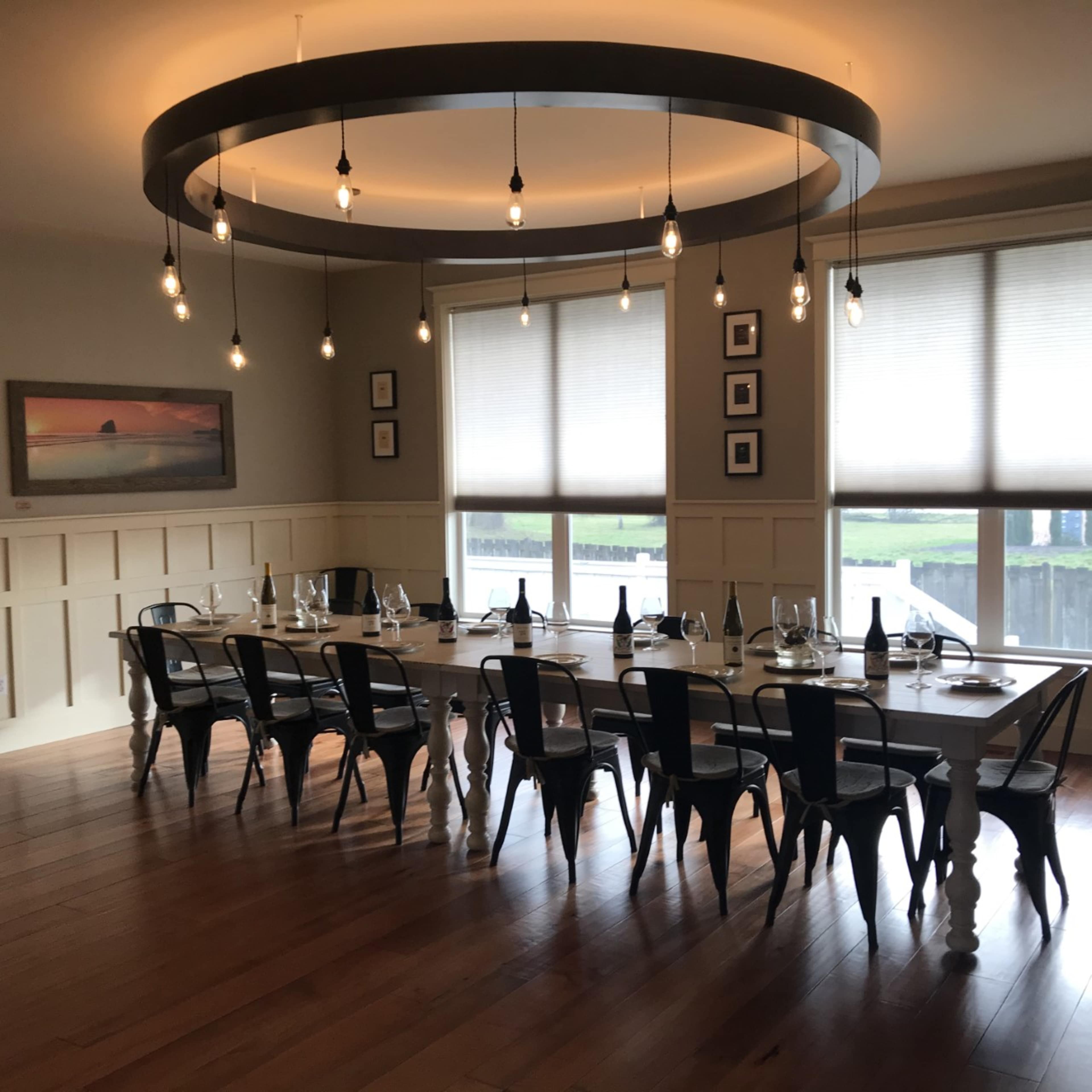 A long dining table is set with wine glasses and bottles beneath a circular light fixture in a well-lit room with large windows.