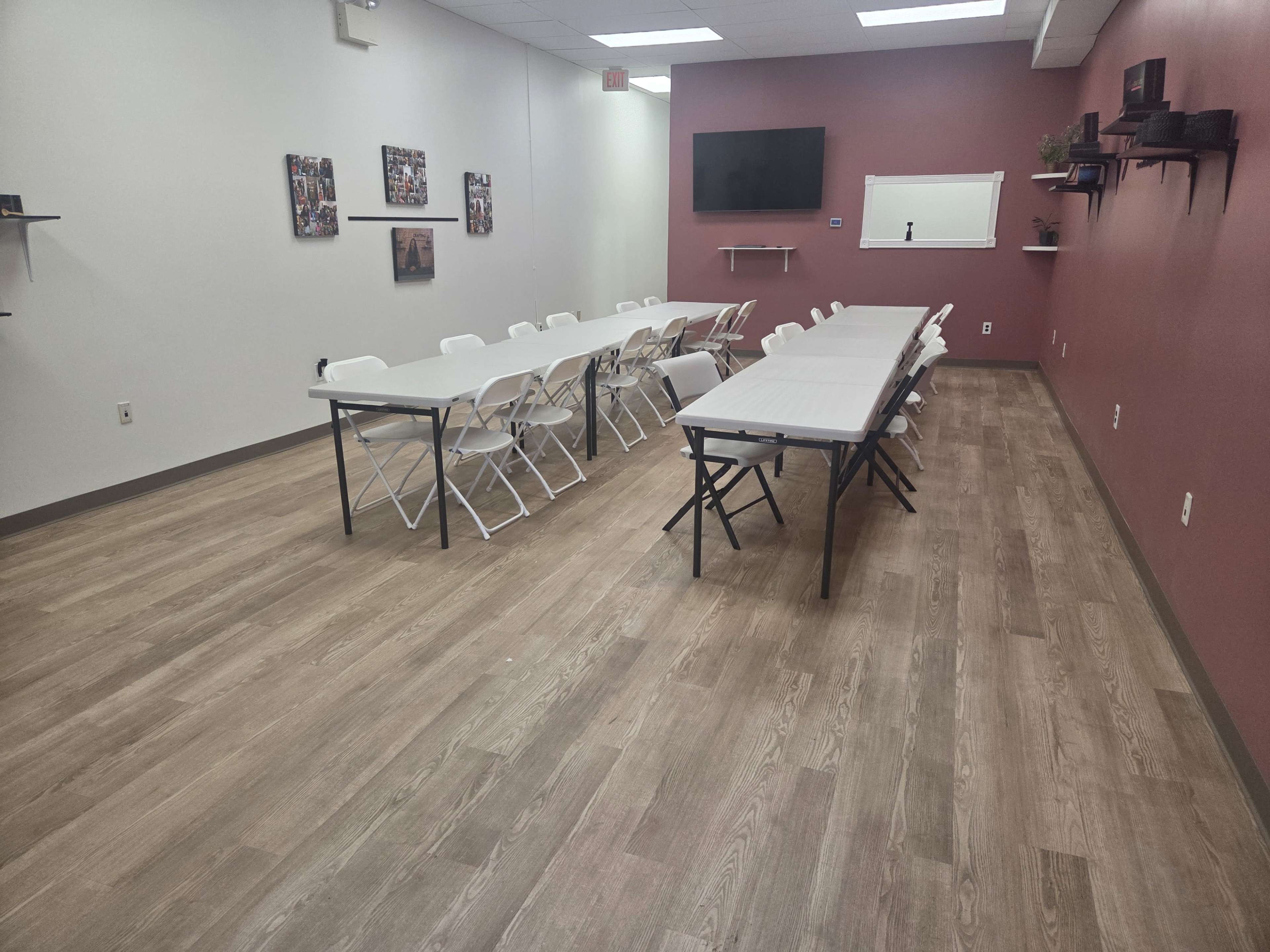 The image shows a sparsely furnished meeting room with a long table and chairs arranged in two lines against a red and white wall.